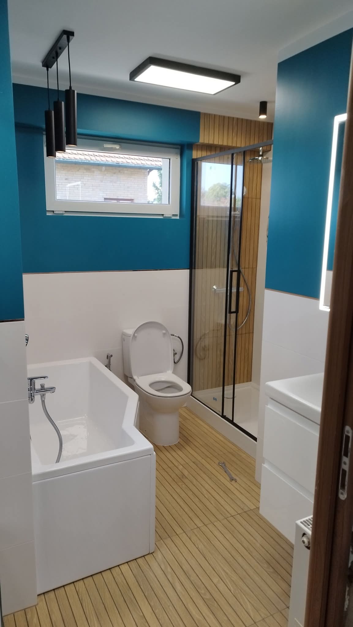 Nowoczesna łazienka z turkusowymi ścianami, drewnianą podłogą i kabiną prysznicową z czarnymi ramami. Nad wanną wiszą trzy czarne lampy.