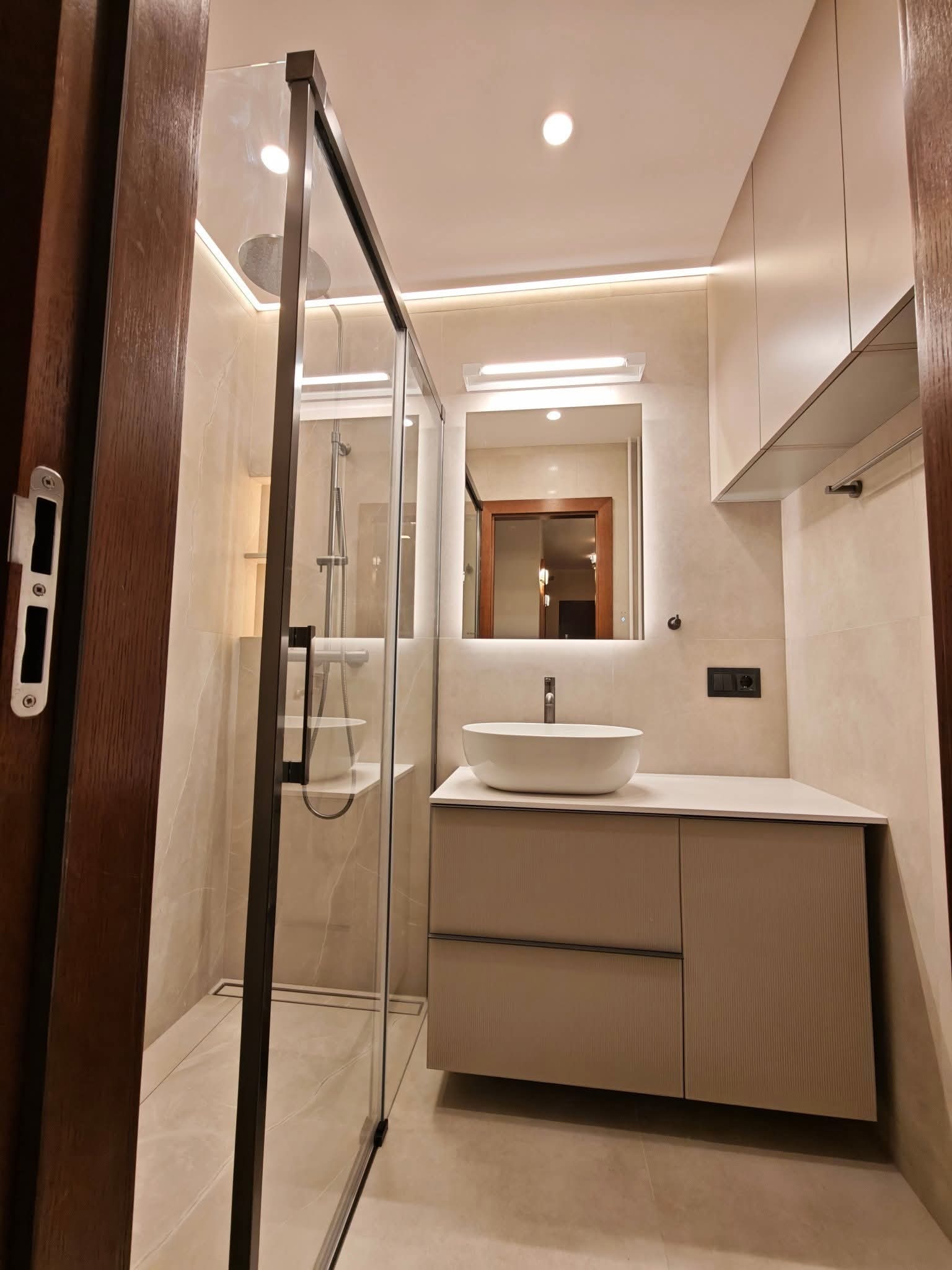 Nowoczesna łazienka w beżach: kabina prysznicowa ze szklaną ścianką, umywalka nablatowa na szafce z frezowanymi frontami, lustro z podświetleniem LED i subtelne oświetlenie.