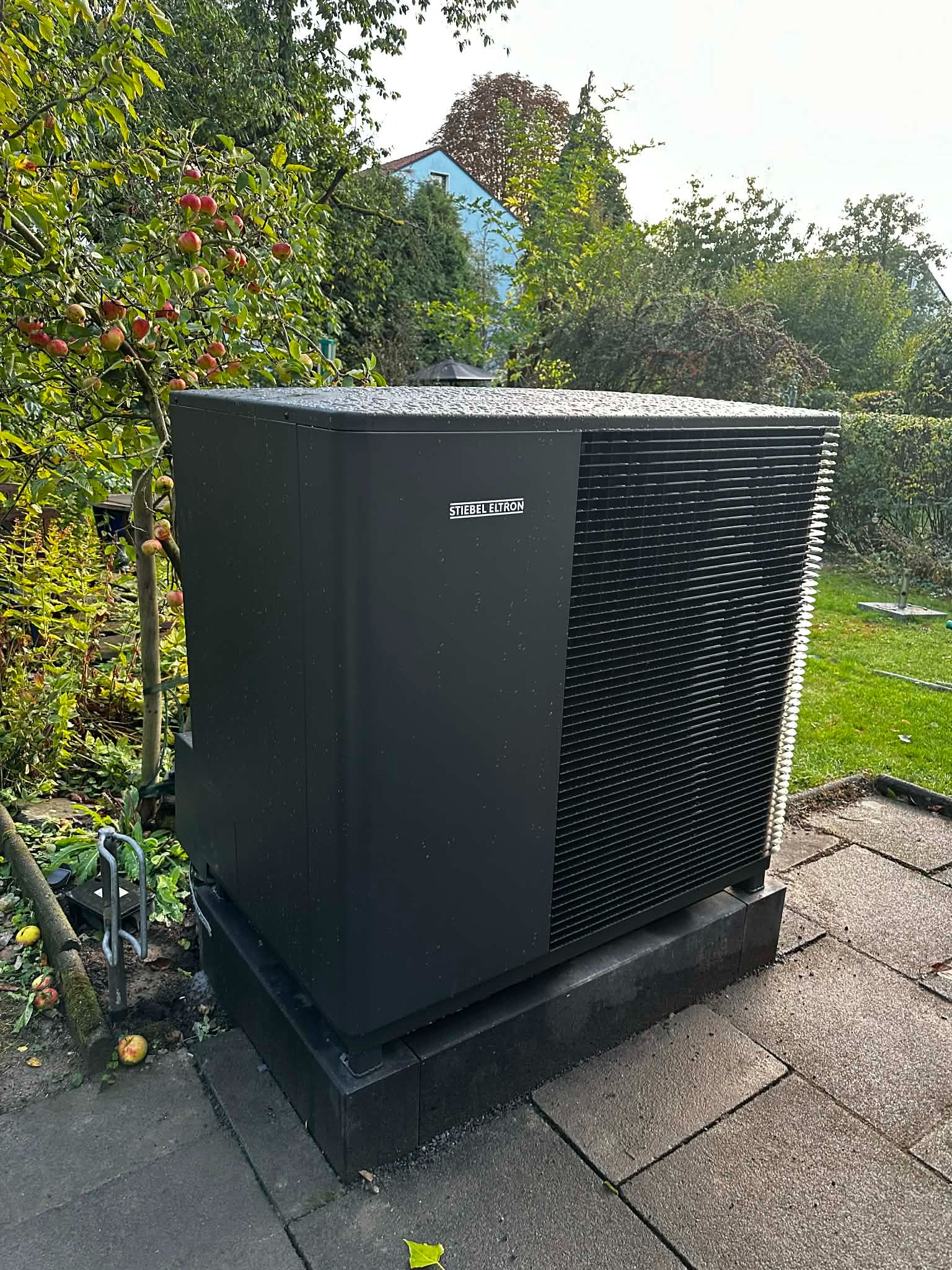 Czarna pompa ciepła Stibel Eltron na zewnątrz, otoczona zielenią i drzewem z jabłkami. Widoczne krople deszczu na obudowie. Tło stanowi dom i zieleń.