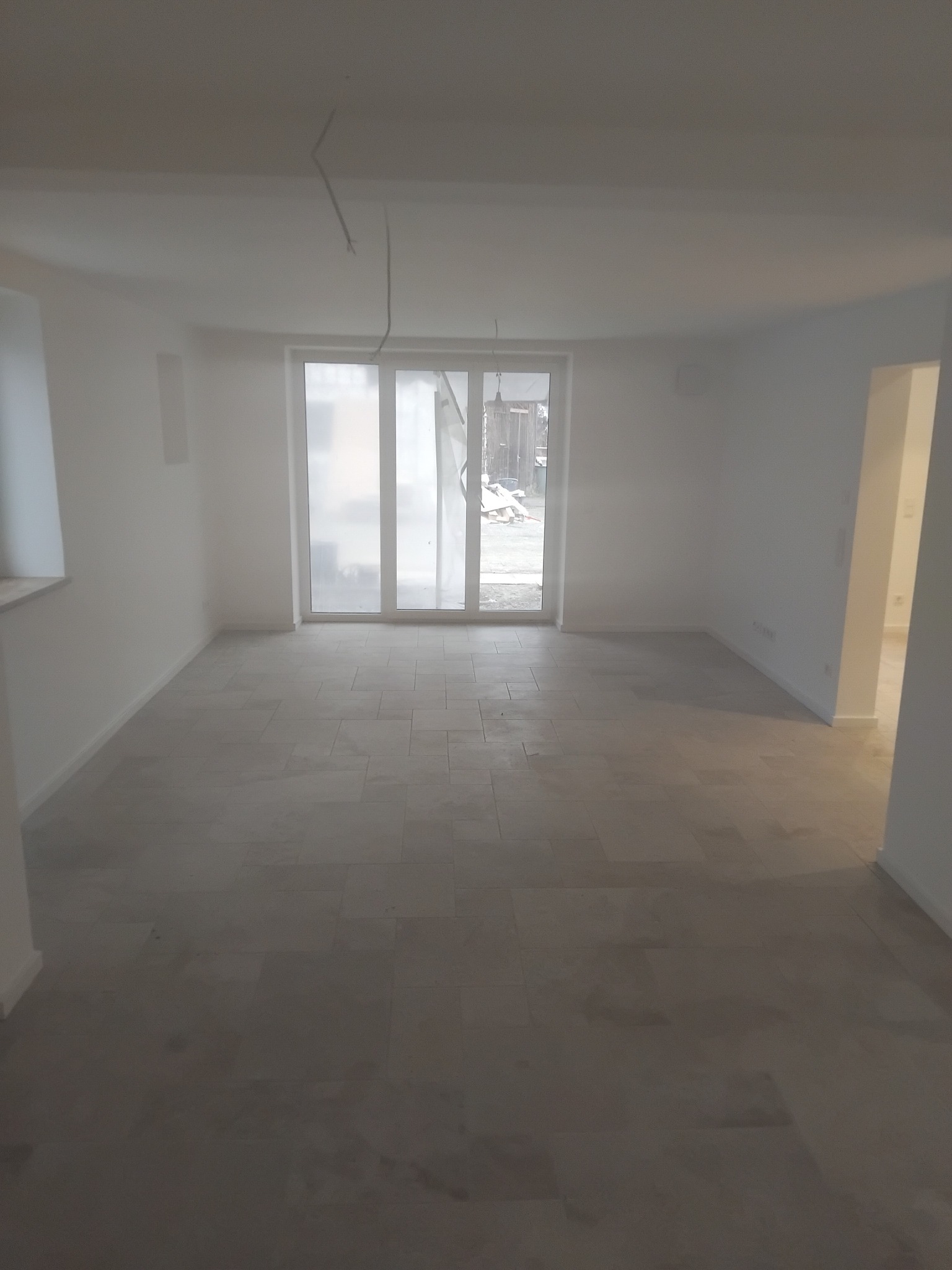 Jasne wnętrze po remoncie z ułożoną podłogą z płytek, widoczne białe ściany i duże okno balkonowe. Na suficie widoczne kable.