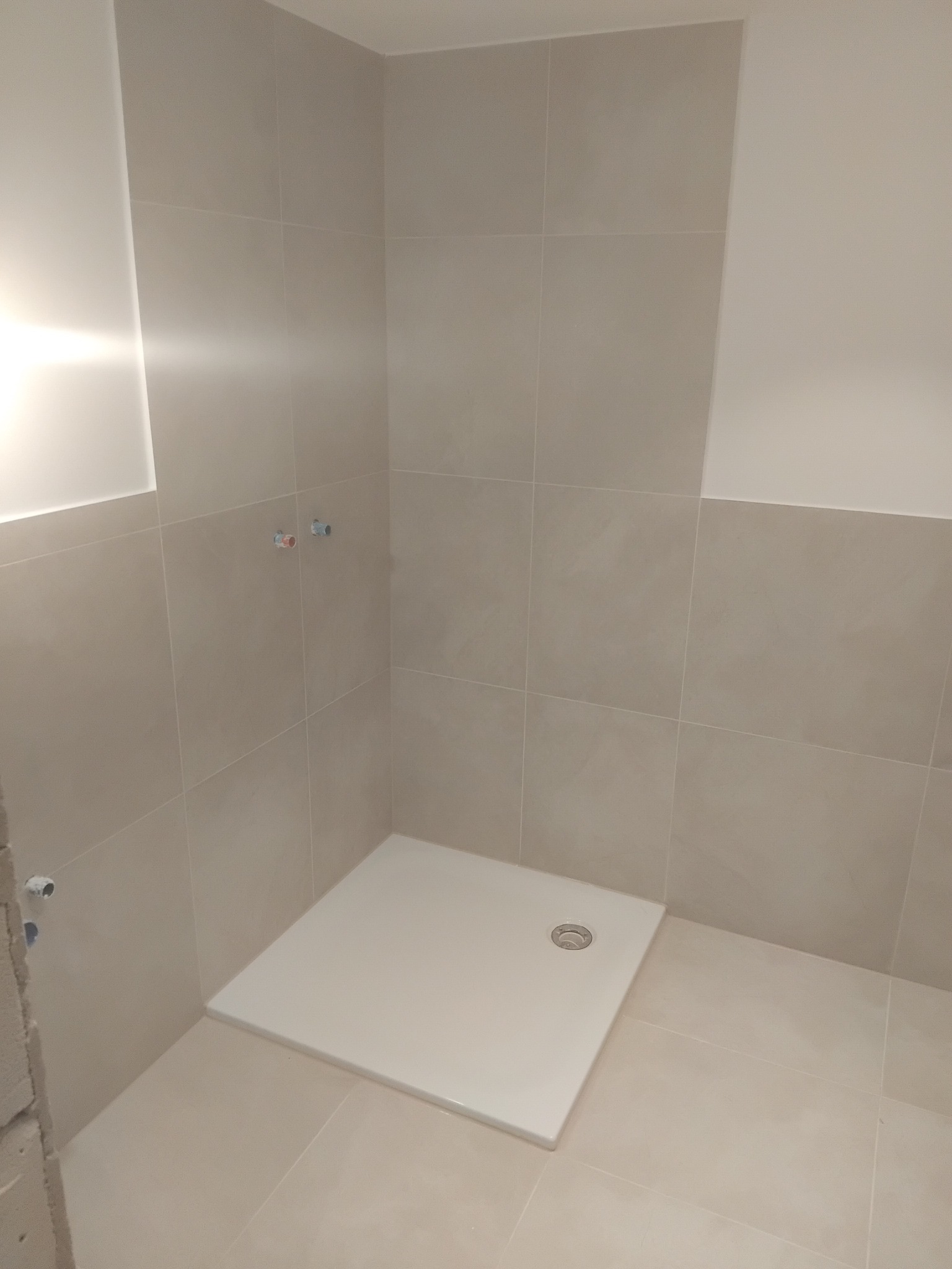 Wykończona kabina prysznicowa z beżowymi płytkami, widoczne przyłącza wodne. Minimalistyczny design, jasne wnętrze. Ukończony etap prac w łazience.