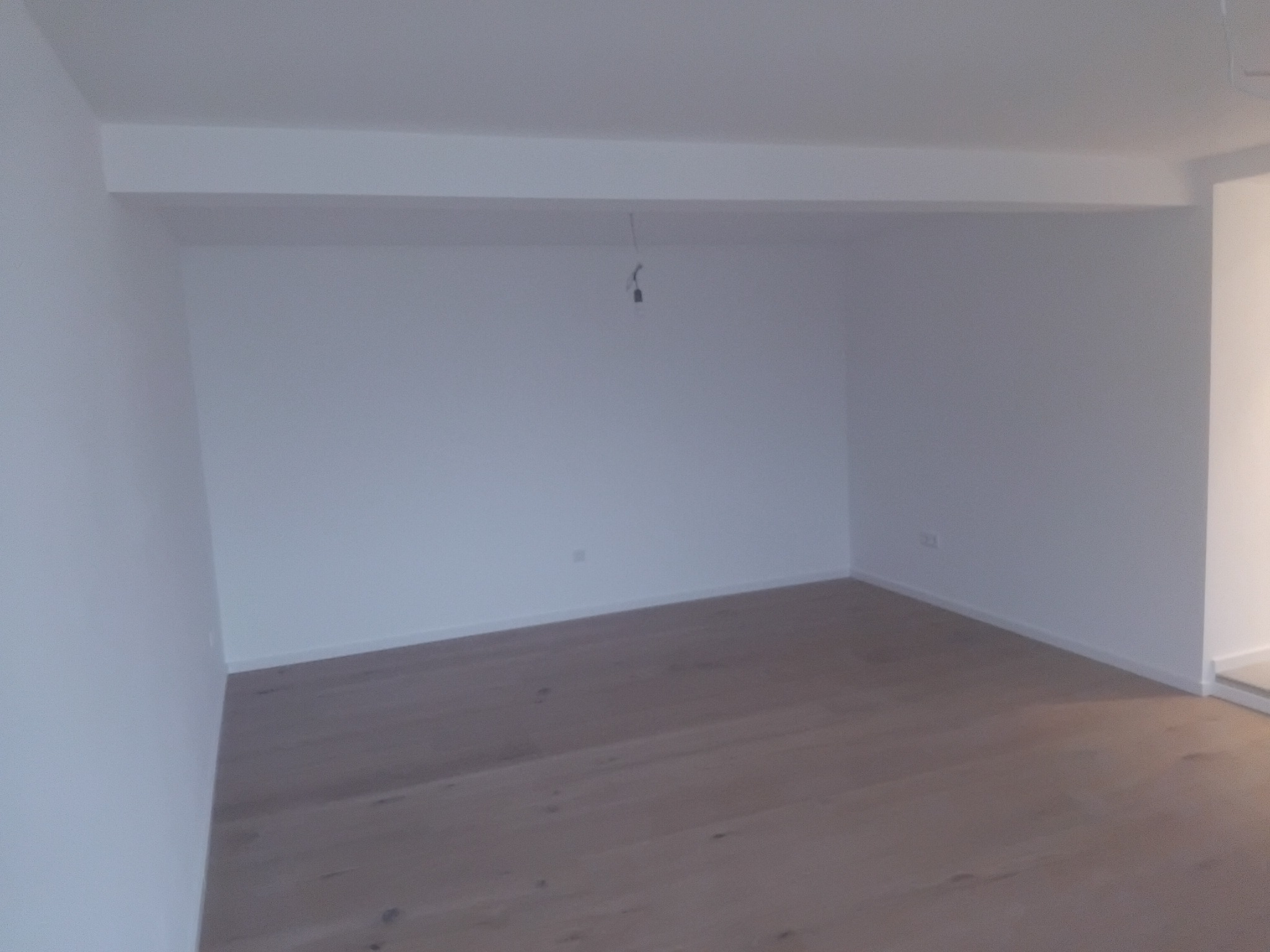 Jasne, drewniane panele podłogowe w pustym, świeżo pomalowanym pokoju z białymi ścianami. Widoczny kabel zwisający z sufitu.