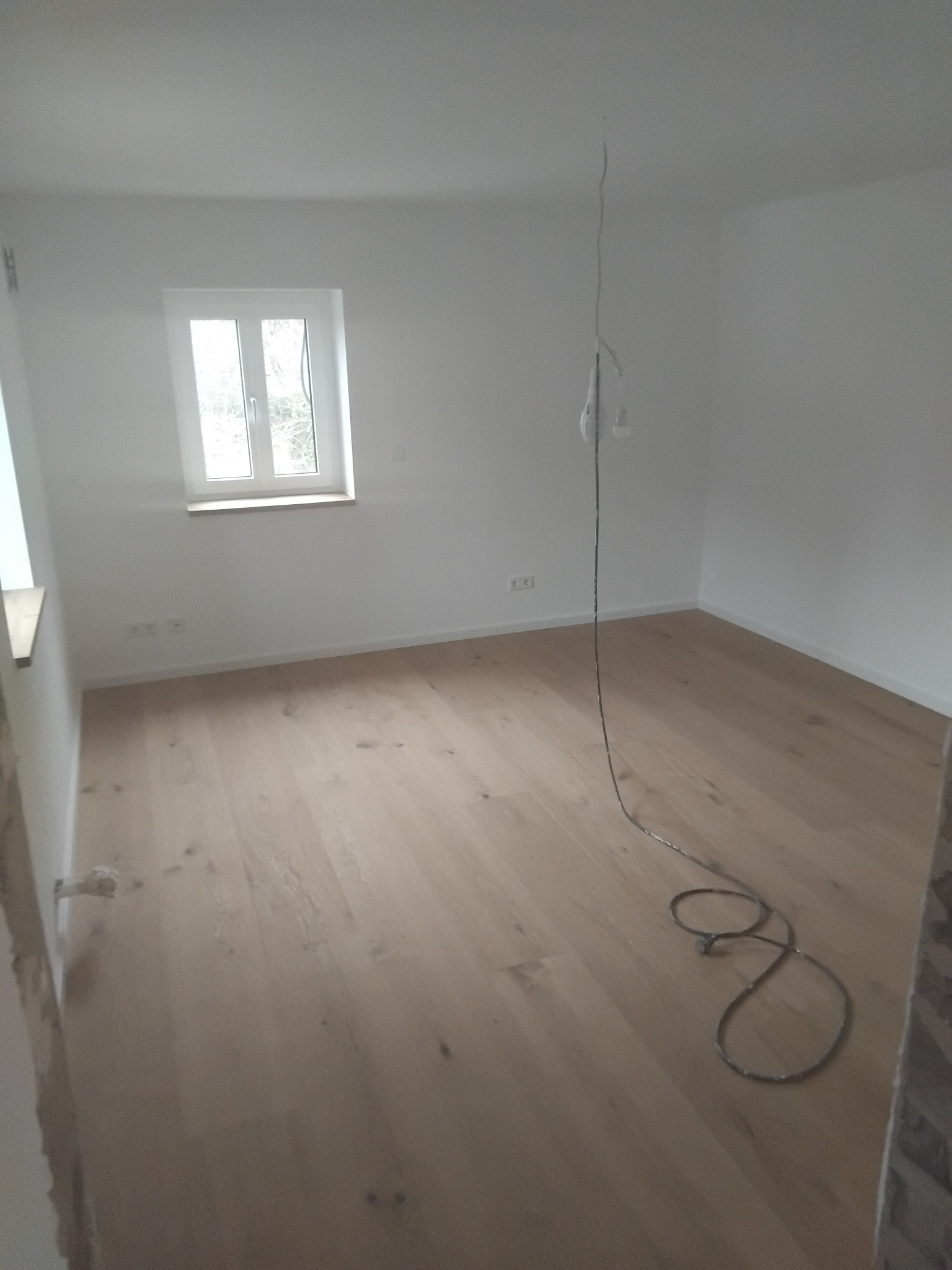 Jasny, drewniany parkiet w pokoju z białymi ścianami i oknem. Kabel z żarówką zwisa z sufitu. Widoczne gniazdka elektryczne.