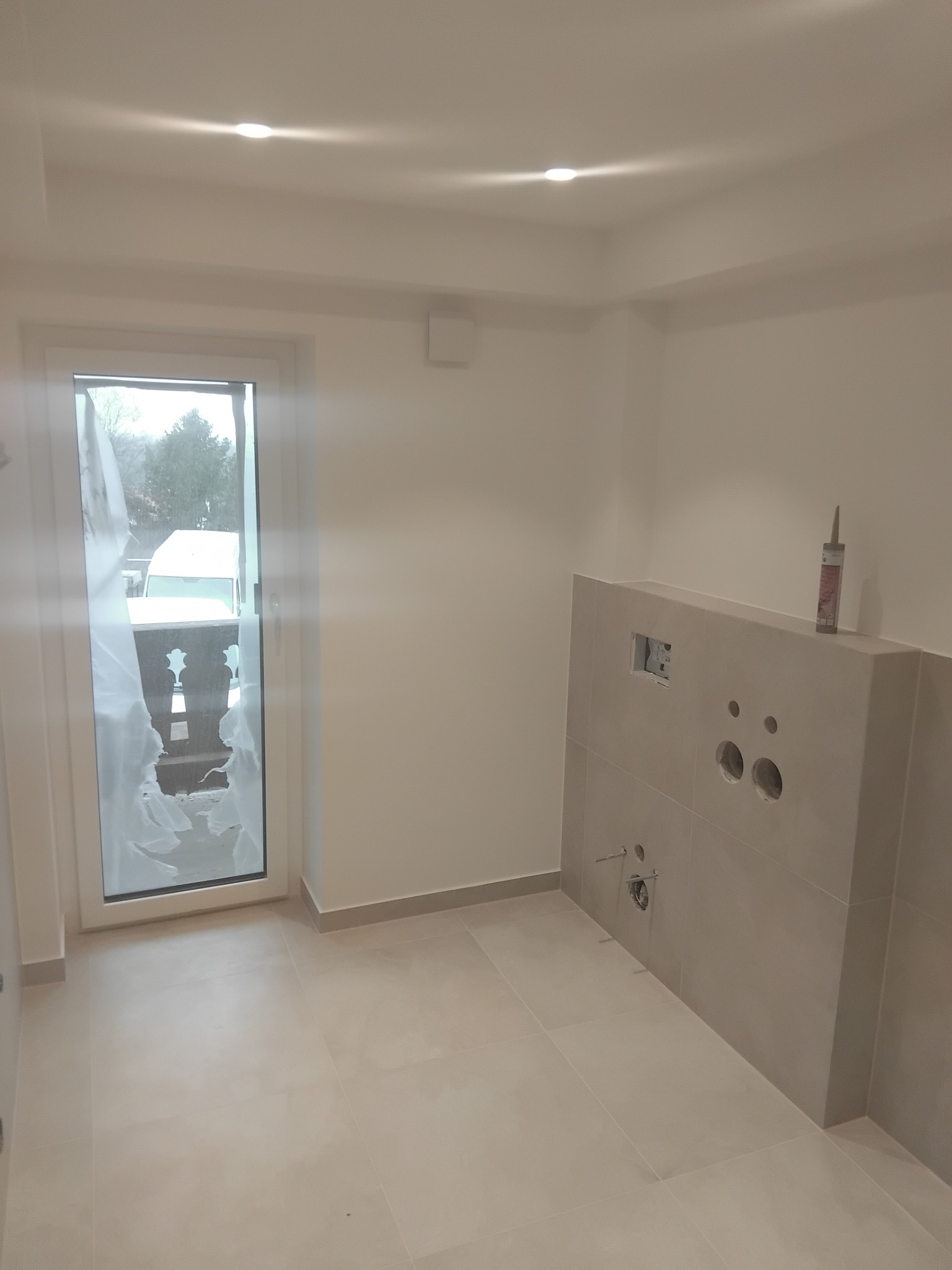 Jasna łazienka w trakcie wykańczania: beżowe płytki, przygotowane otwory na armaturę, okno balkonowe z widokiem na zewnątrz. Tubka silikonu na ścianie.