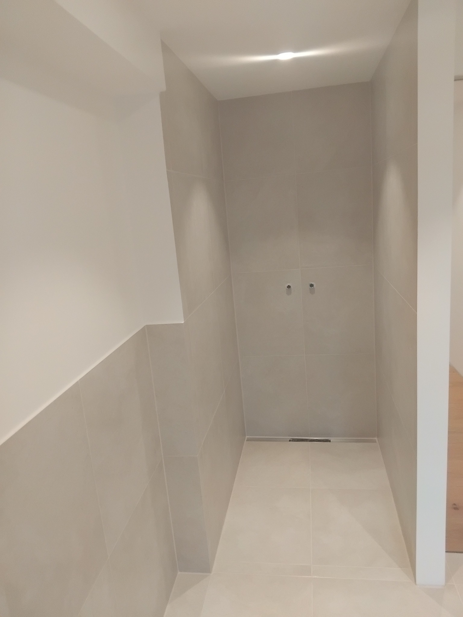 Wykończona kabina prysznicowa z beżowymi płytkami, widoczny odpływ liniowy i przygotowane punkty podłączenia armatury. Minimalistyczny design, oświetlenie punktowe.