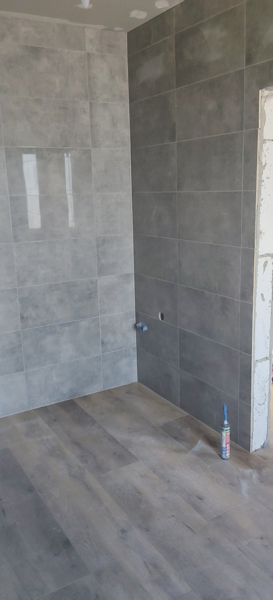 Wykończona łazienka z szarymi płytkami na ścianach i panelami podłogowymi imitującymi drewno. Widoczna tuba silikonu przy ścianie.