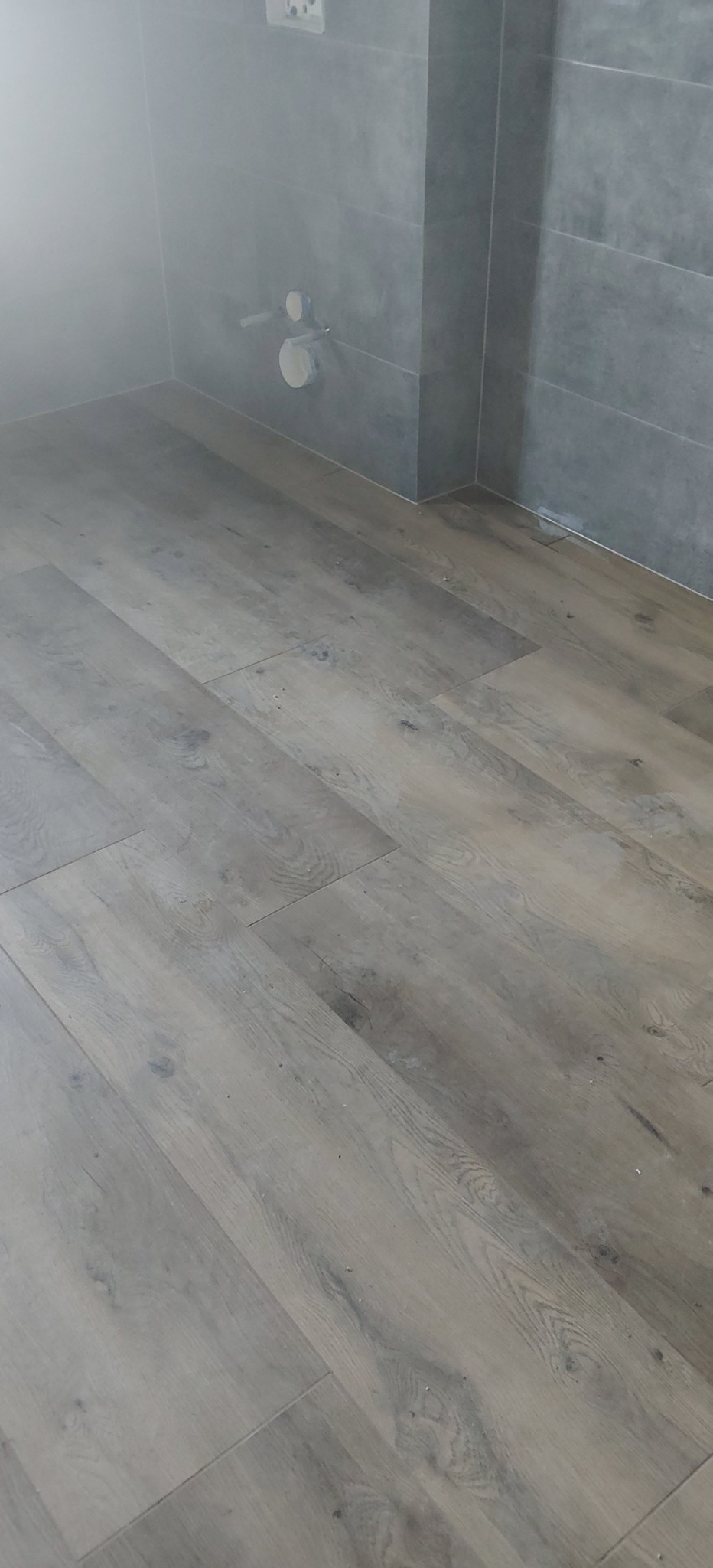 Szare panele podłogowe w łazience z szarą glazurą na ścianach i widoczną instalacją sanitarną. Minimalistyczne wykończenie wnętrza.