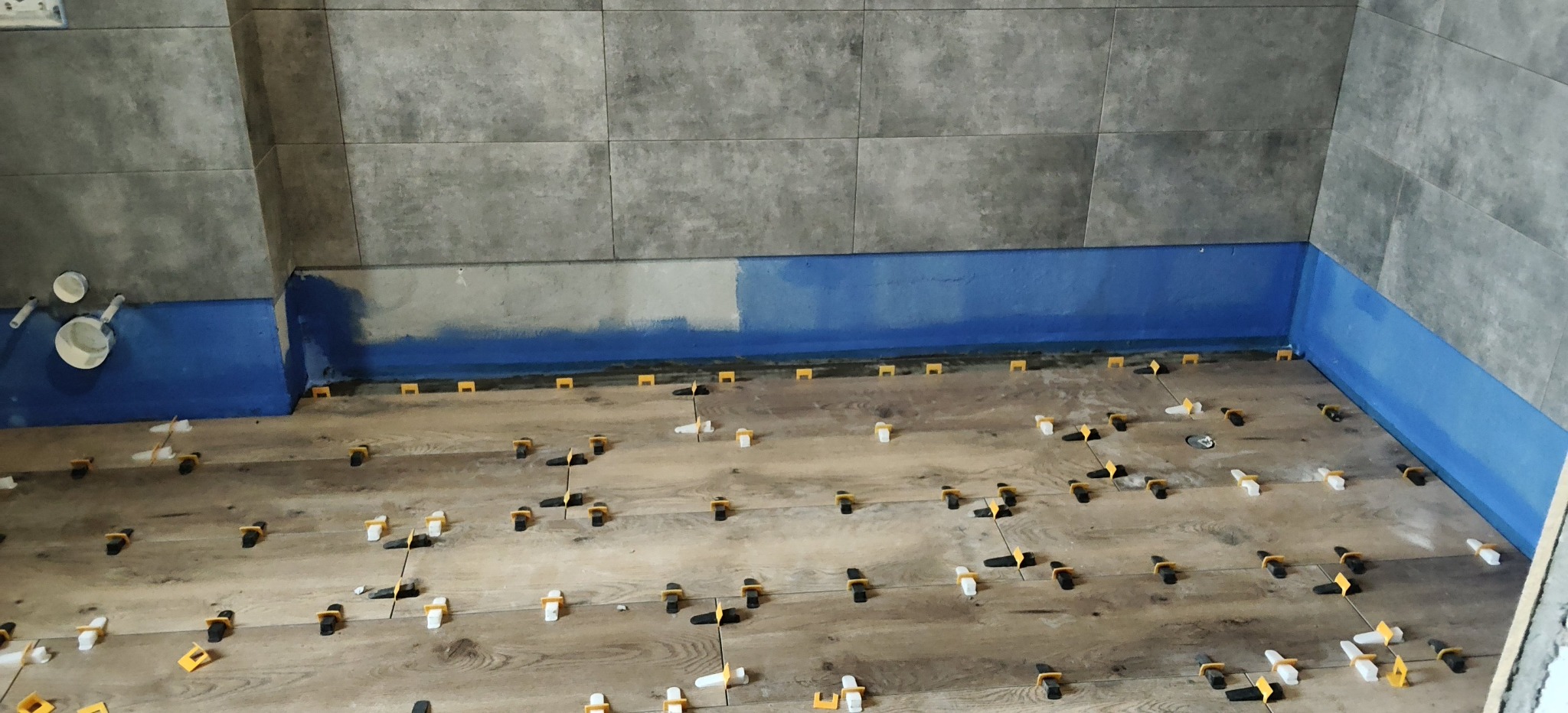 Układanie płytek imitujących drewno na podłodze w łazience, z użyciem systemu poziomowania. Ściany wyłożone szarymi płytkami, zabezpieczone niebieską folią w płynie.