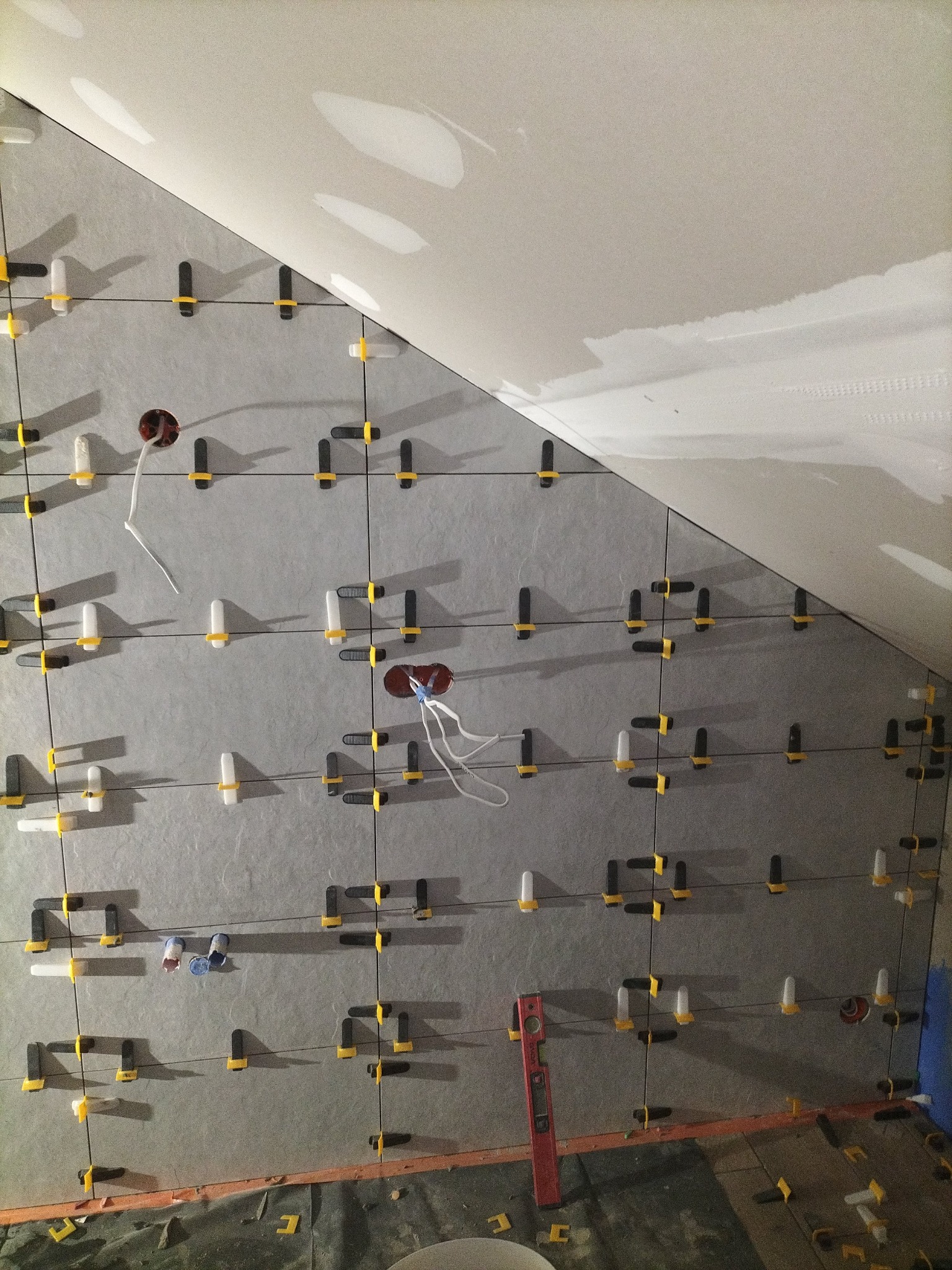 Ściana w trakcie układania szarych płytek z użyciem systemu poziomowania; widoczne otwory na instalacje i poziomica przy krawędzi. Ukośny sufit w tle.