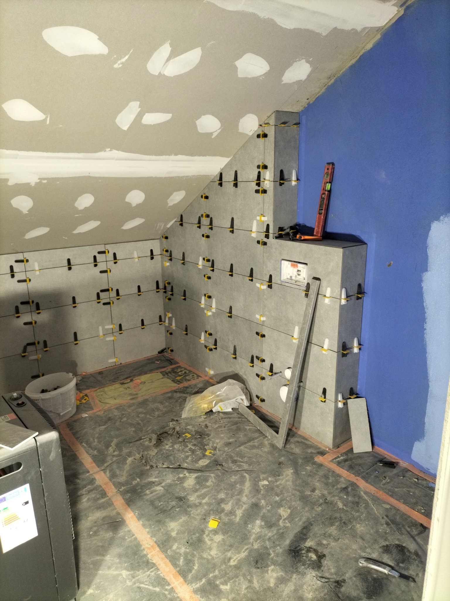 Układanie szarych płytek w łazience na poddaszu, z użyciem poziomicy i klipsów poziomujących. Ściany częściowo pomalowane na niebiesko, sufit z płyt karton-gips.