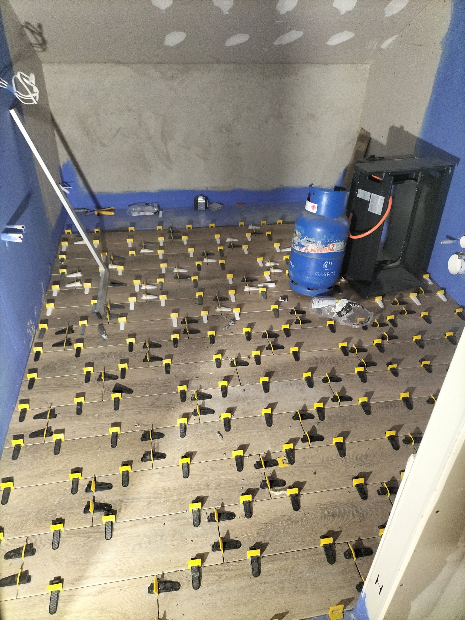 Układanie drewnopodobnych płytek podłogowych z użyciem systemu poziomowania; widoczna butla gazowa i narzędzia glazurnicze w surowym wnętrzu.