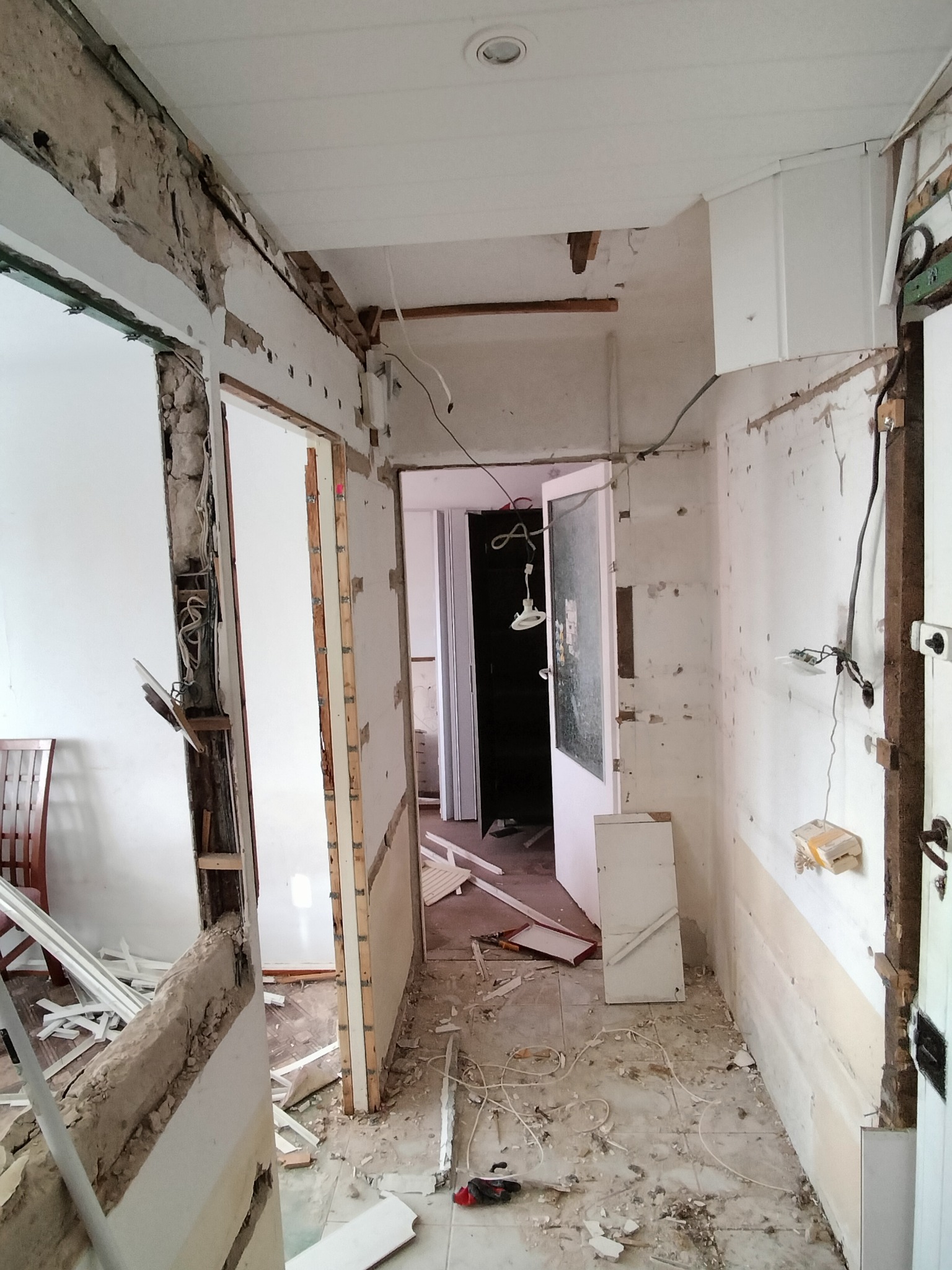 Wnętrze w trakcie demontażu: zerwane tapety, odsłonięte kable, gruz na podłodze. Widok na korytarz z otwartymi drzwiami. Etap przygotowawczy do remontu.