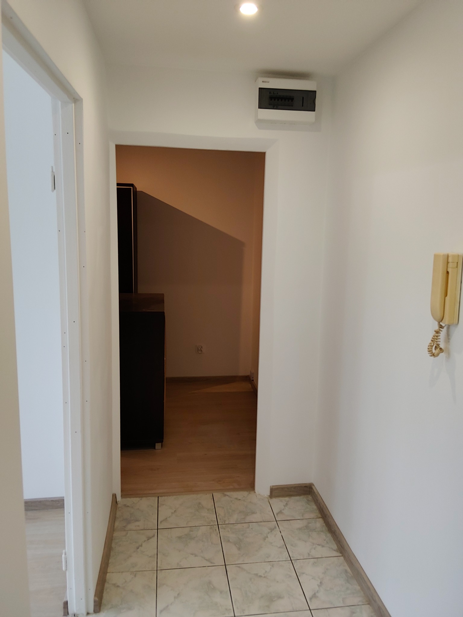 Jasne wnętrze po odświeżeniu: białe ściany, płytki podłogowe w odcieniach beżu i szarości, widoczna skrzynka elektryczna i domofon. Minimalistyczny design.