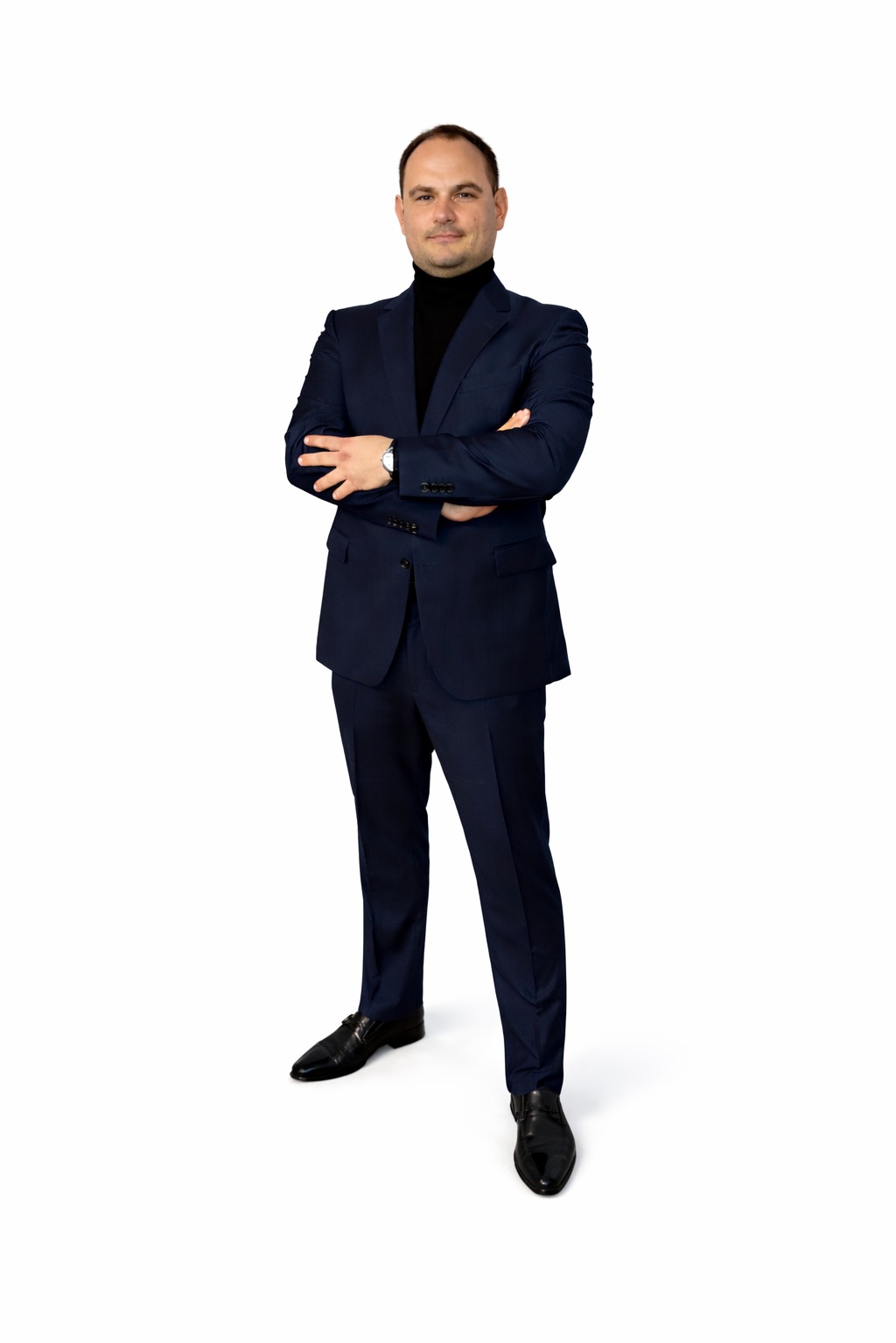 Mężczyzna w granatowym garniturze i czarnym golfie, stoi na białym tle z założonymi rękami, prezentując profesjonalny wizerunek.