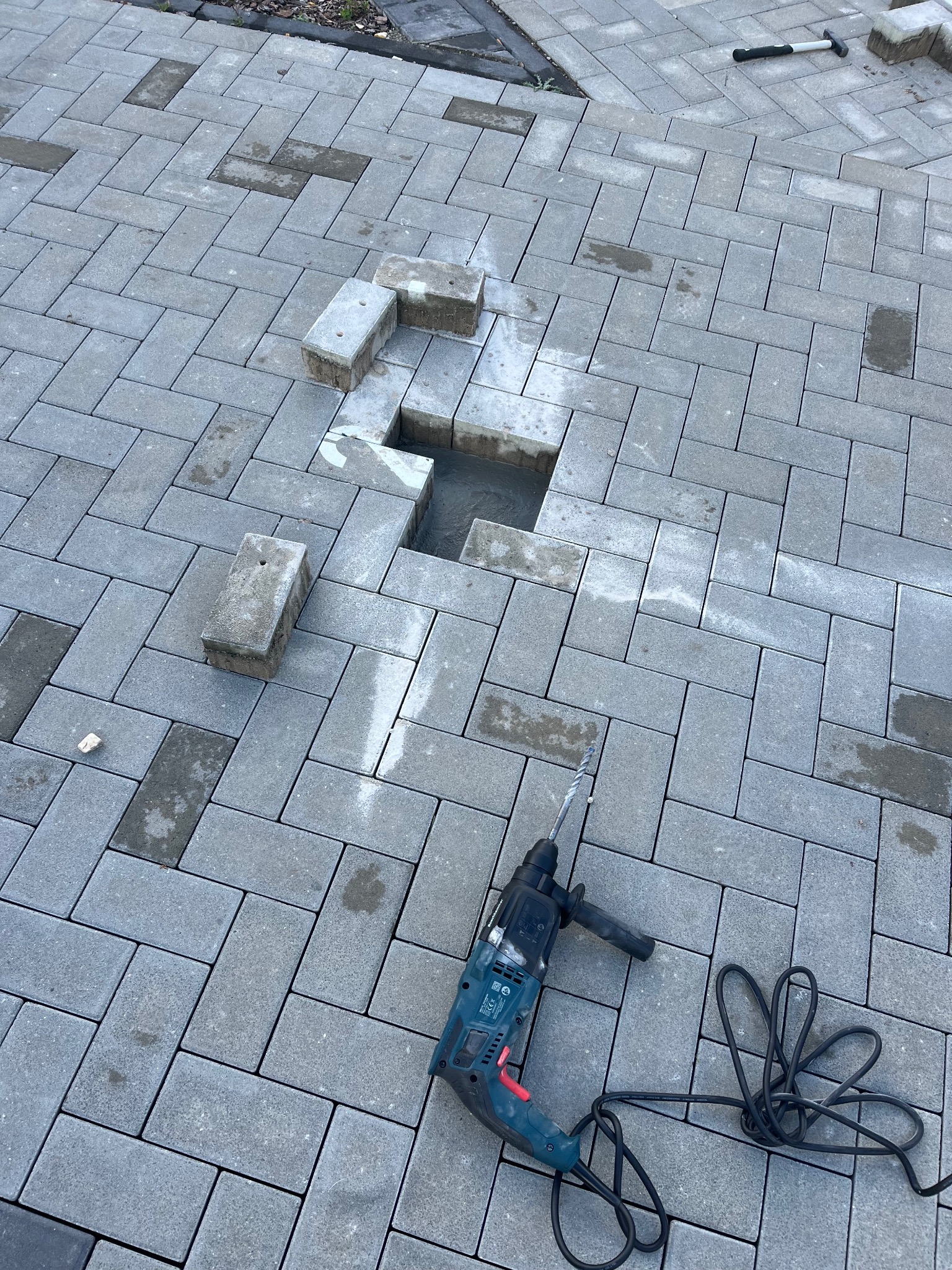Układanie szarej kostki brukowej w jodełkę, z widocznym otworem i leżącymi obok kostkami. W kadrze wiertarka udarowa z przewodem. Praca w toku.