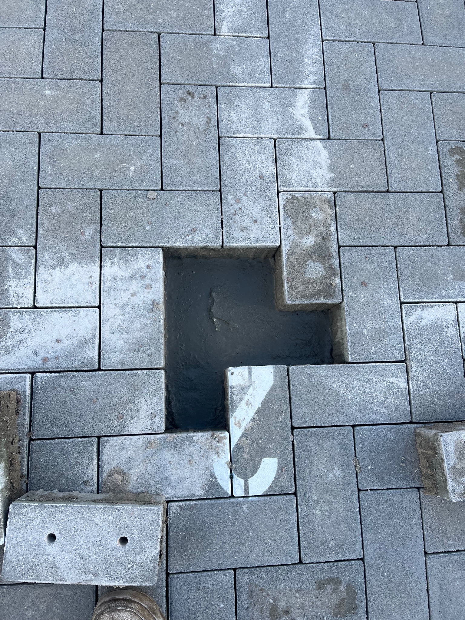 Szara kostka brukowa ułożona w regularny wzór, z widocznym otworem po usuniętej kostce i fragmentem buta w rogu kadru. Widoczne ślady cementu.