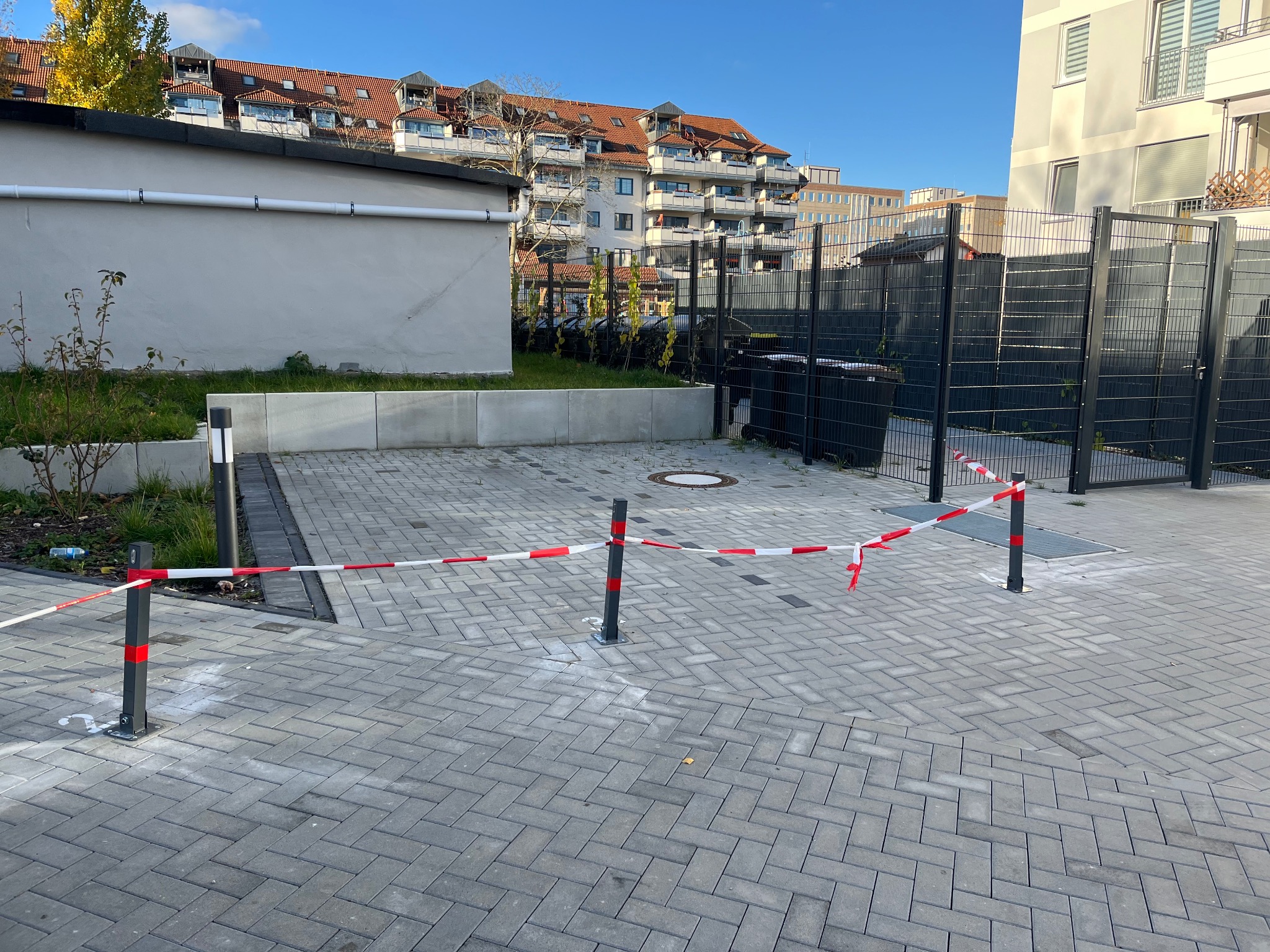 Szara kostka brukowa ułożona w jodełkę, zabezpieczona czerwono-białą taśmą na słupkach. W tle budynki mieszkalne i fragment ogrodzenia.