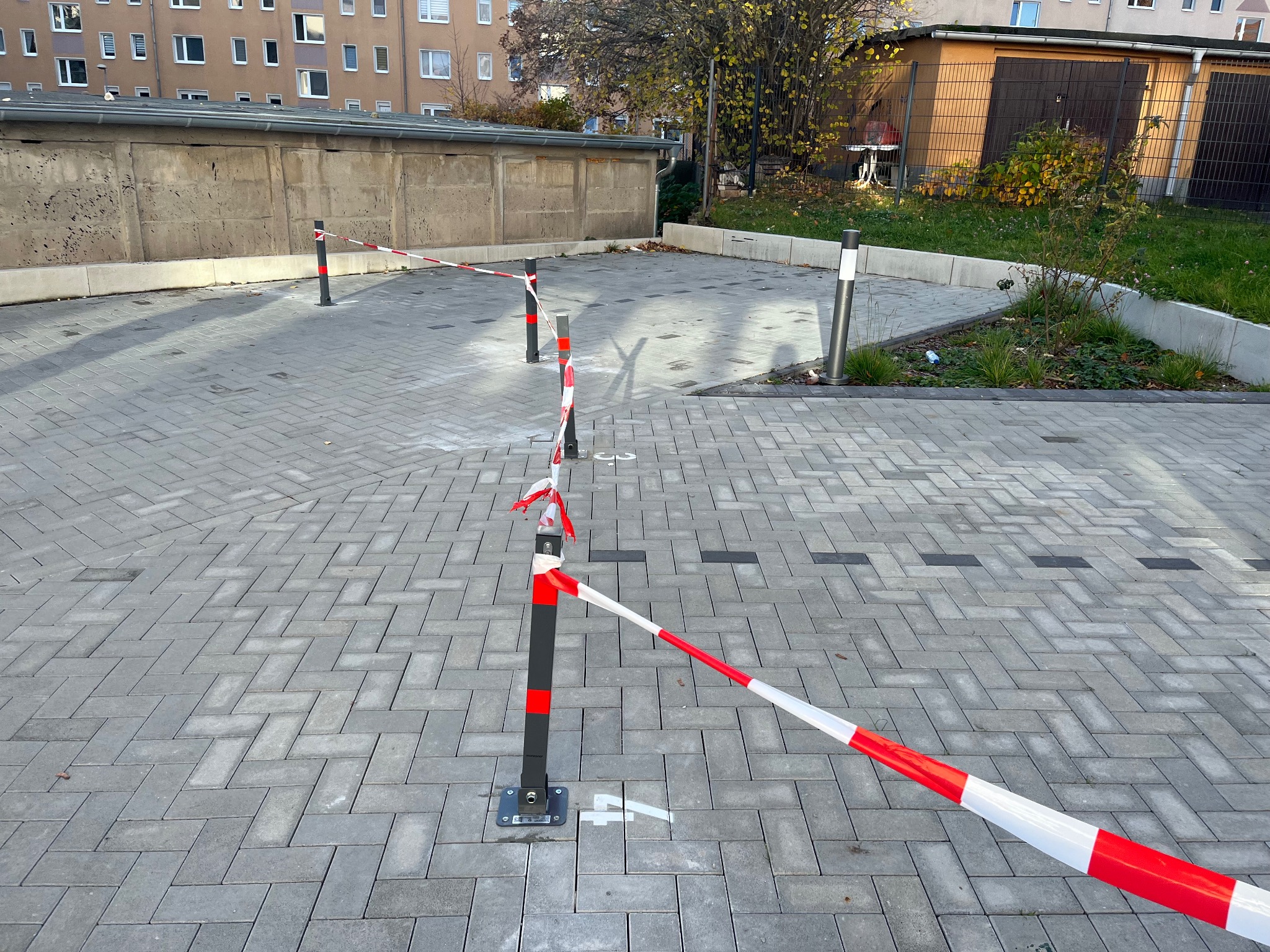 Szara kostka brukowa z wyznaczonymi słupkami i taśmą ostrzegawczą na parkingu. Widoczne budynki w tle. Oznakowanie terenu po zakończonych pracach.