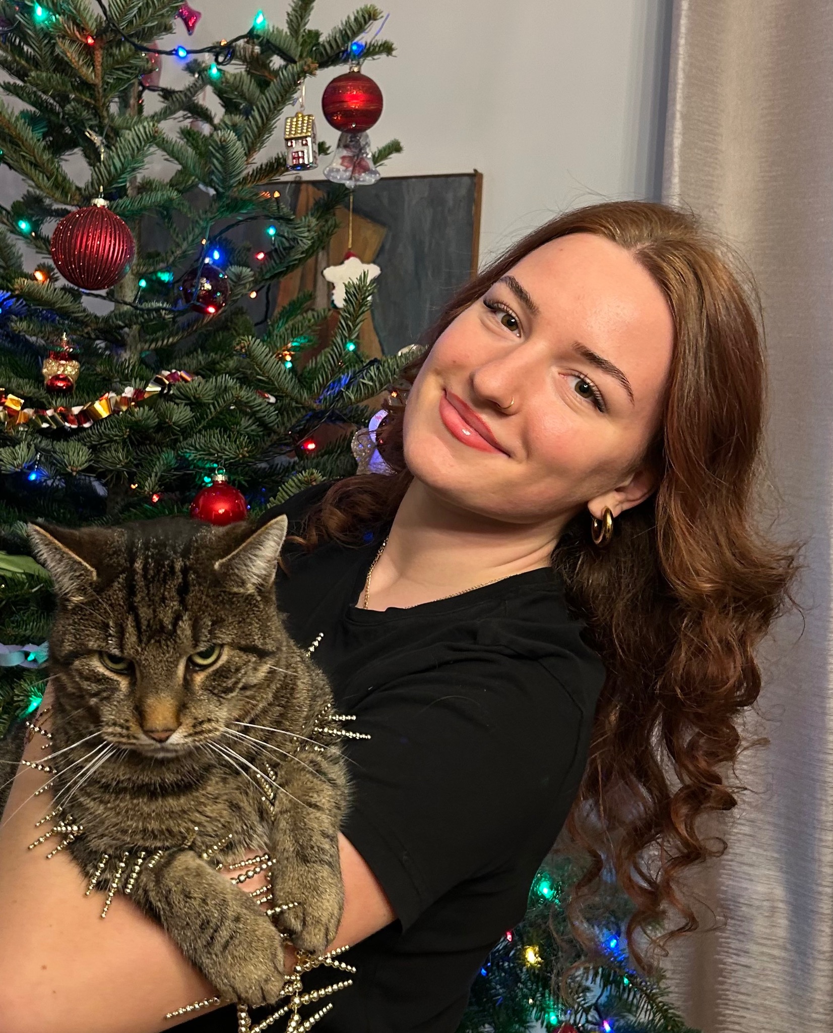 Uśmiechnięta kobieta z rudymi włosami trzyma kota w ozdobnej obroży na tle choinki z lampkami i bombkami. Portret w świątecznym klimacie.