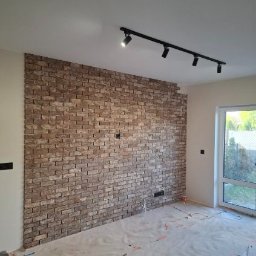 Ruslan Andrushchyshyn - Ściana wykończona cegłą dekoracyjną w jasnych odcieniach brązu, oświetlona czarną listwą z reflektorami. Podłoga zabezpieczona folią malarską.