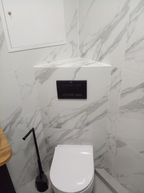 Biała toaleta wisząca z czarnym przyciskiem spłukującym na tle marmurowych płytek. Widoczny fragment blatu i szczotka do WC. Wykończenie łazienki w nowoczesnym stylu.