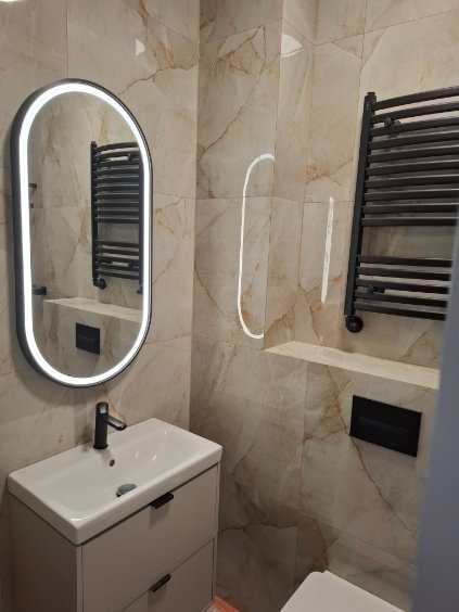 Wykończona łazienka z beżowymi płytkami imitującymi marmur, owalnym lustrem z podświetleniem LED, umywalką z szafką i grzejnikiem drabinkowym.