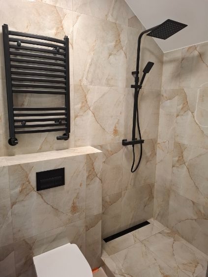 Wykończona łazienka z beżowymi płytkami imitującymi marmur, czarną armaturą (deszczownica, grzejnik) i odpływem liniowym. Minimalistyczny styl, nowoczesne rozwiązania.