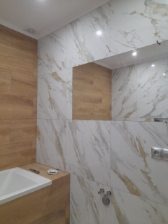 Biało-złota glazura imitująca marmur na ścianach łazienki, z akcentem drewnopodobnych płytek i lustrem. Widoczna wanna i bateria. Oświetlenie punktowe.