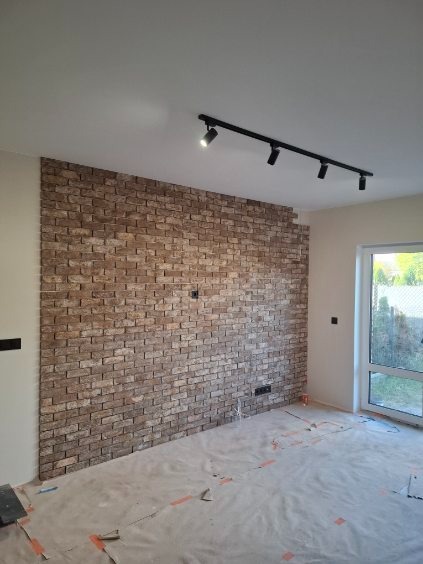 Ściana wykończona cegłą dekoracyjną w jasnych odcieniach brązu, oświetlona czarną listwą z reflektorami. Podłoga zabezpieczona folią malarską.