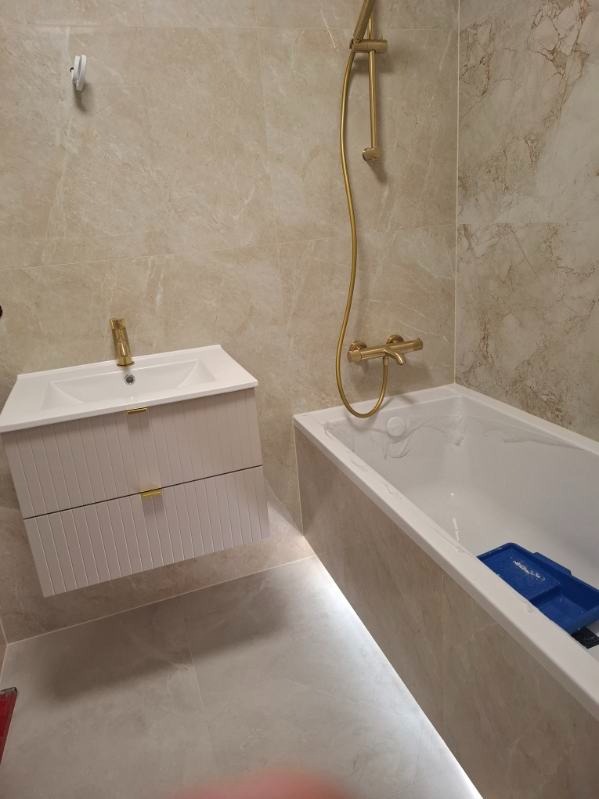 Jasna łazienka z beżowymi płytkami imitującymi marmur. Biała szafka pod umywalką z złotą armaturą i bateria prysznicowa w tym samym kolorze. Podświetlana wanna.
