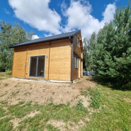 Cichoracki budowlany Świat Maciej Cichoracki - Nowoczesny, drewniany domek letniskowy z dużymi przesuwnymi drzwiami, na lekkim wzniesieniu, otoczony zielenią i błękitnym niebem z chmurami.