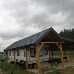 Cichoracki budowlany Świat Maciej Cichoracki - Mobilny dom z szarą blachodachówką i drewnianą konstrukcją zadaszenia tarasu, otoczony zielenią, pod pochmurnym niebem. Widoczne elementy ogrodu i las w tle.