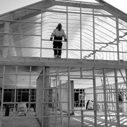 Cichoracki budowlany Świat Maciej Cichoracki - Czarno-białe zdjęcie konstrukcji domu w trakcie budowy, z pracownikiem na górze, ukazujące drewniany szkielet i elementy konstrukcyjne.