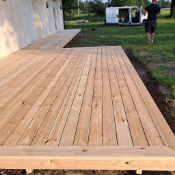 Cichoracki budowlany Świat Maciej Cichoracki - Nowy, drewniany taras z jasnych desek przy domu z białymi roletami, z widocznym fragmentem ogrodu i samochodem dostawczym w tle. Widok z bliska na deski.