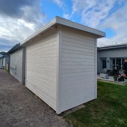 Cichoracki budowlany Świat Maciej Cichoracki - Biała, drewniana altana z płaskim dachem, widoczna z boku na tle pochmurnego nieba i zielonego trawnika. Minimalistyczny design, poziome deski.