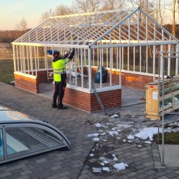 Cichoracki budowlany Świat Maciej Cichoracki - Montaż szklarni z białym szkieletem na ceglanej podmurówce. Pracownik w odblaskowej kamizelce mocuje elementy konstrukcji. W tle drzewa i błękitne niebo.