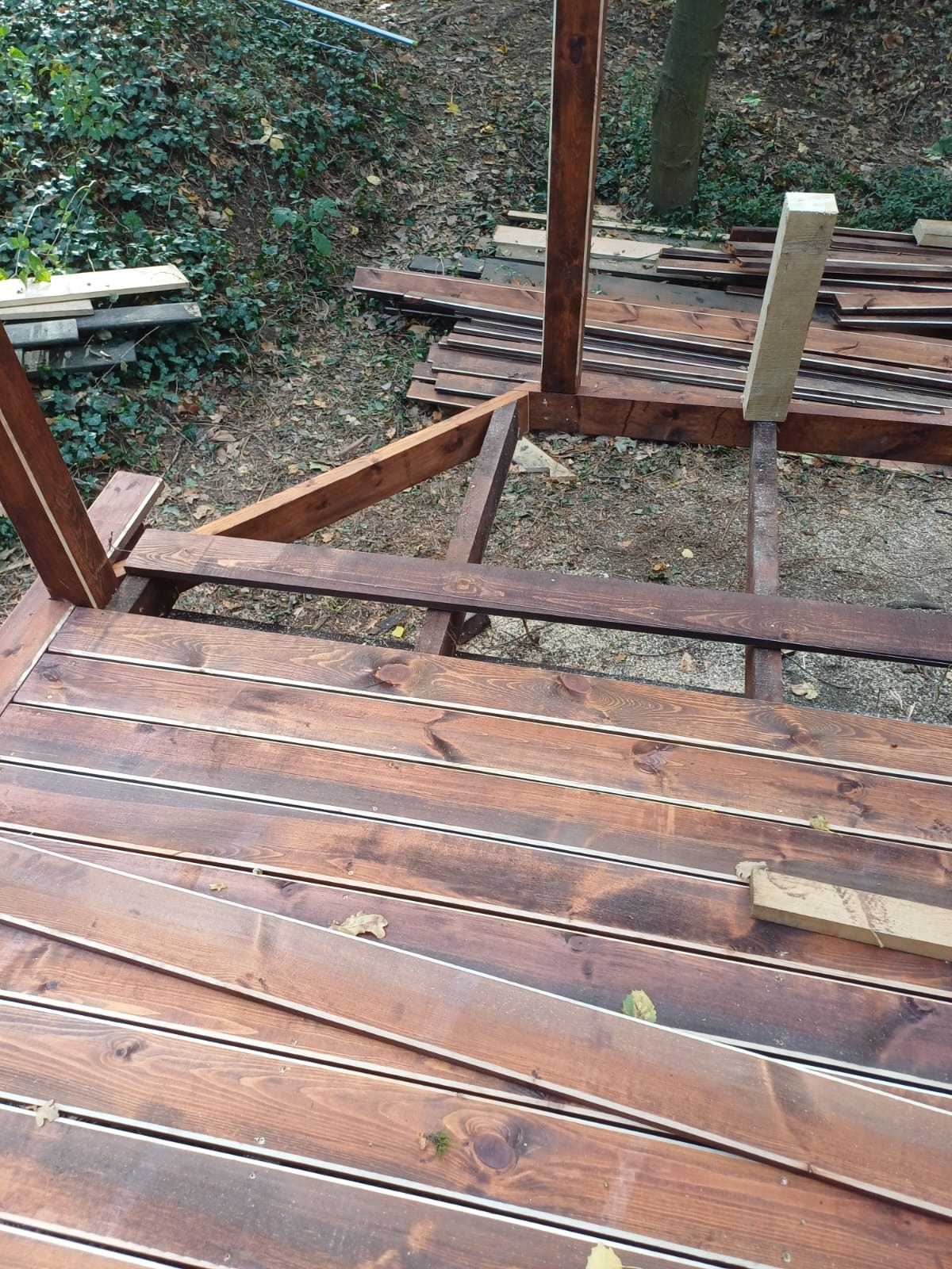 Konstrukcja tarasu drewnianego w trakcie budowy, widoczne deski podłogowe i elementy szkieletu, z zapasem materiału obok. Ujęcie z góry, w naturalnym oświetleniu.