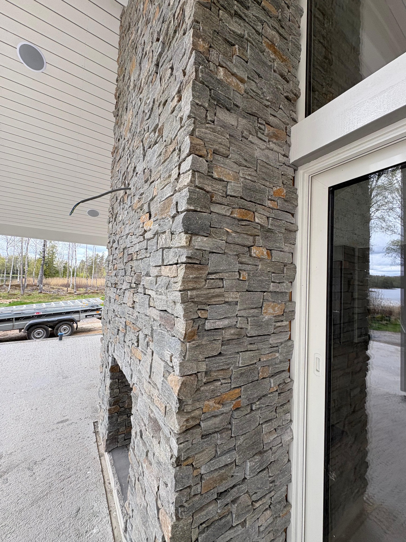 Kamienna elewacja zewnętrzna domu z widocznym fragmentem okna i zarysem krajobrazu w odbiciu szyby. Widoczny fragment przyczepy na zewnątrz.
