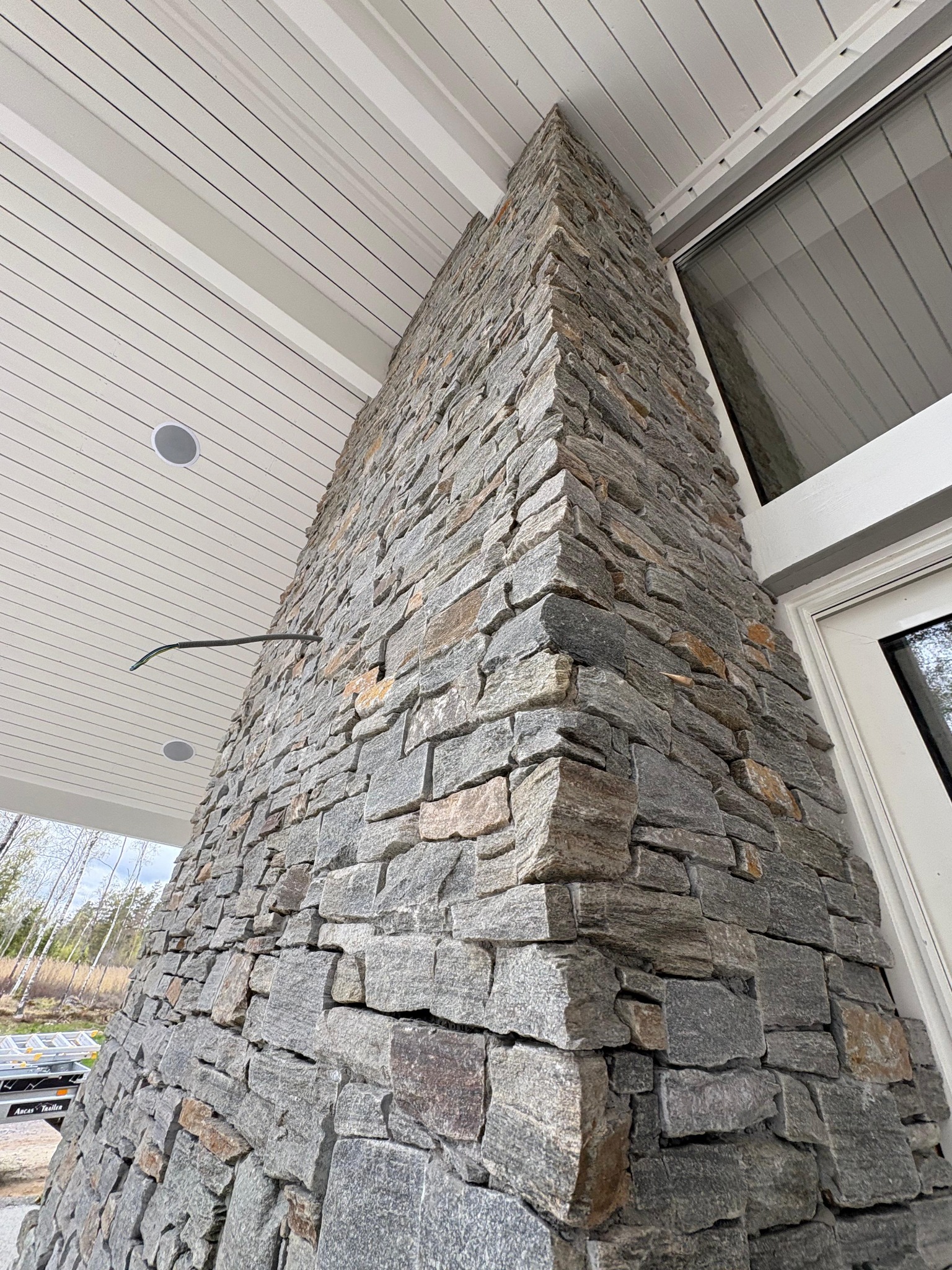 Kamienna elewacja filaru domu z widocznym oknem i podbitką dachową. Surowy kamień w odcieniach szarości i brązu tworzy solidną konstrukcję. Widok z dołu.