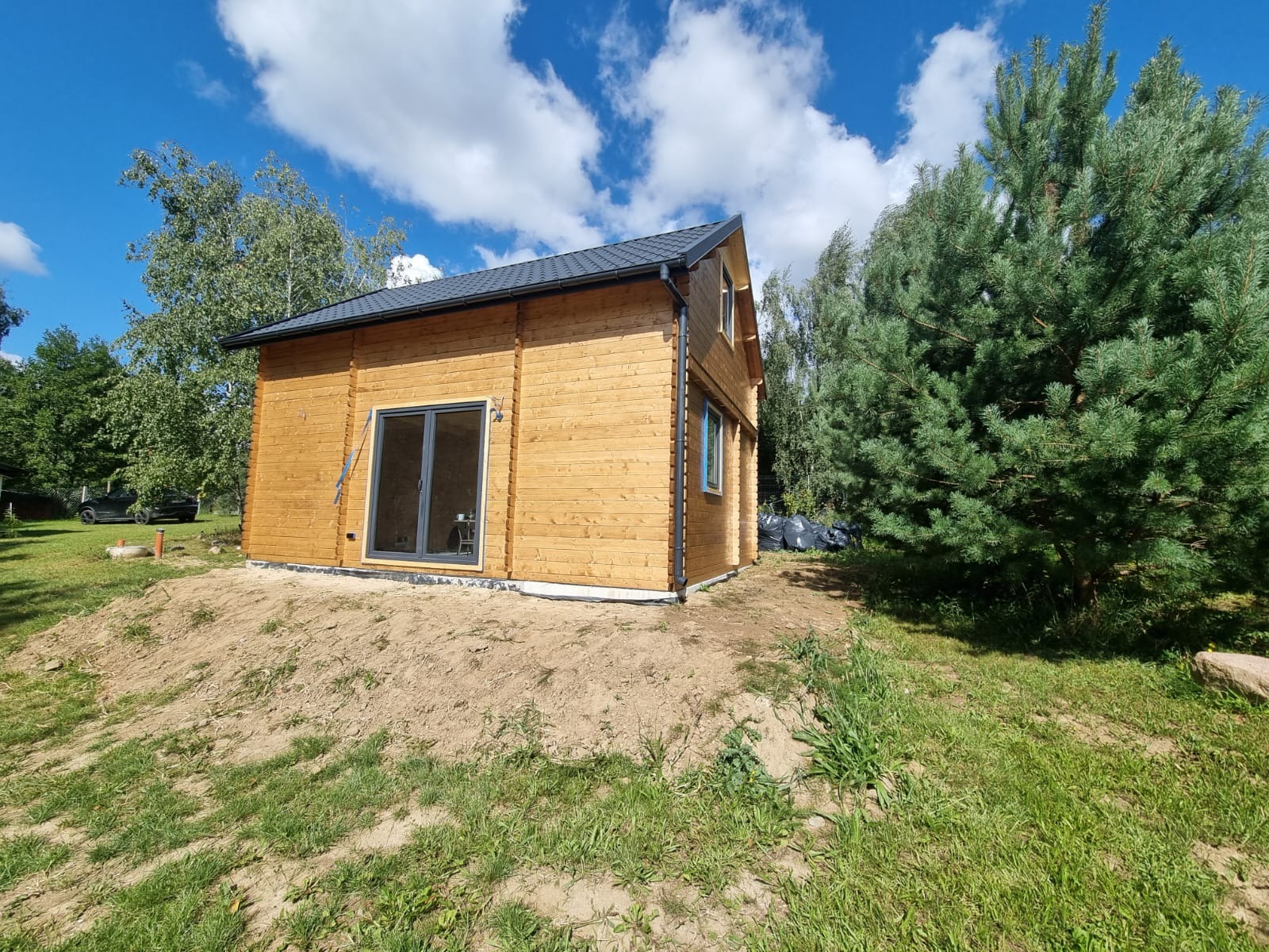 Nowoczesny, drewniany domek letniskowy z dużymi przesuwnymi drzwiami, na lekkim wzniesieniu, otoczony zielenią i błękitnym niebem z chmurami.