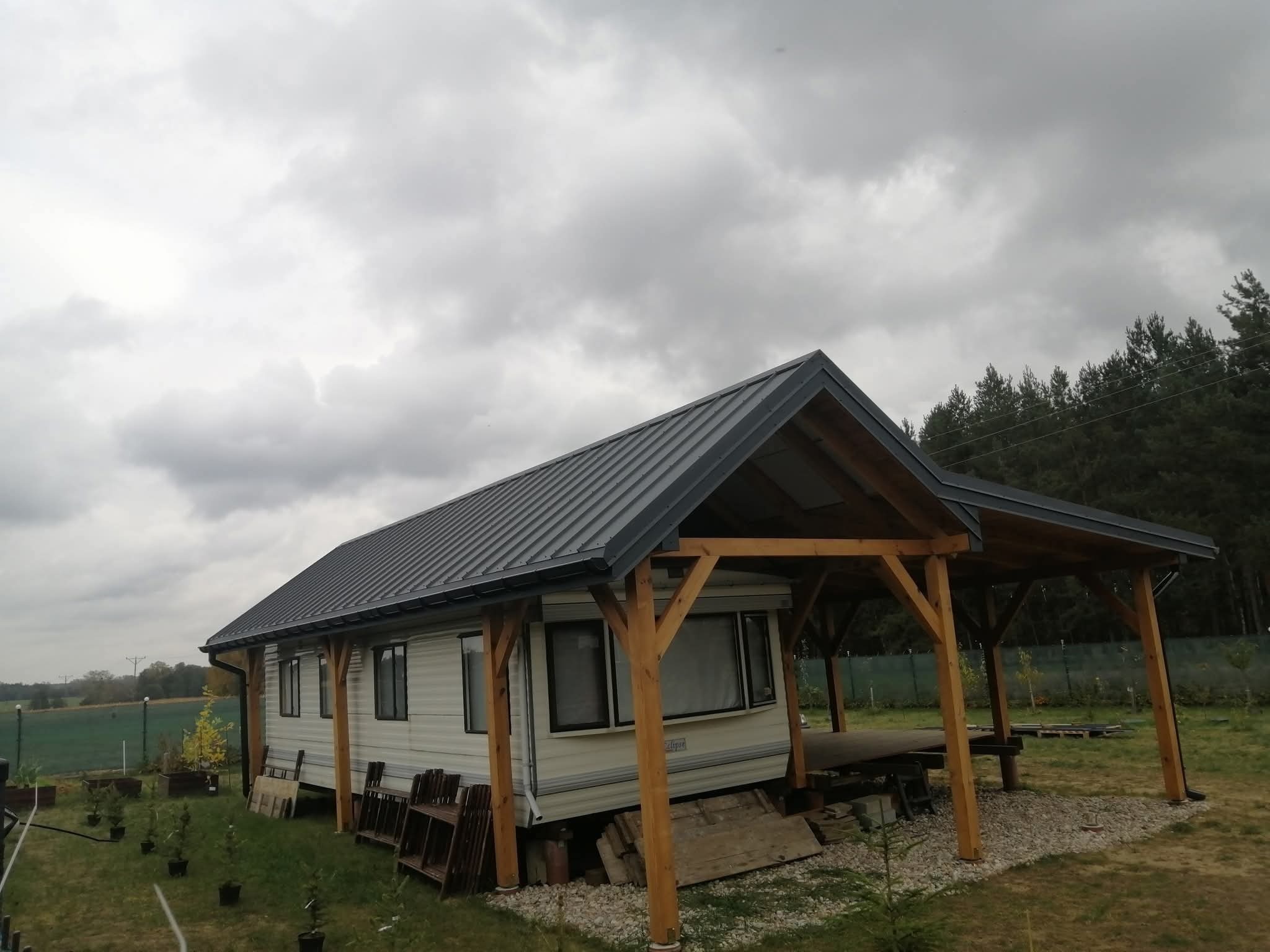 Mobilny dom z szarą blachodachówką i drewnianą konstrukcją zadaszenia tarasu, otoczony zielenią, pod pochmurnym niebem. Widoczne elementy ogrodu i las w tle.