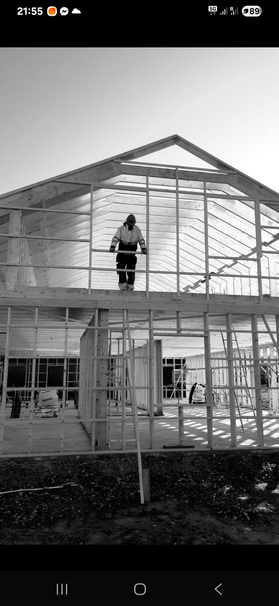 Czarno-białe zdjęcie konstrukcji domu w trakcie budowy, z pracownikiem na górze, ukazujące drewniany szkielet i elementy konstrukcyjne.