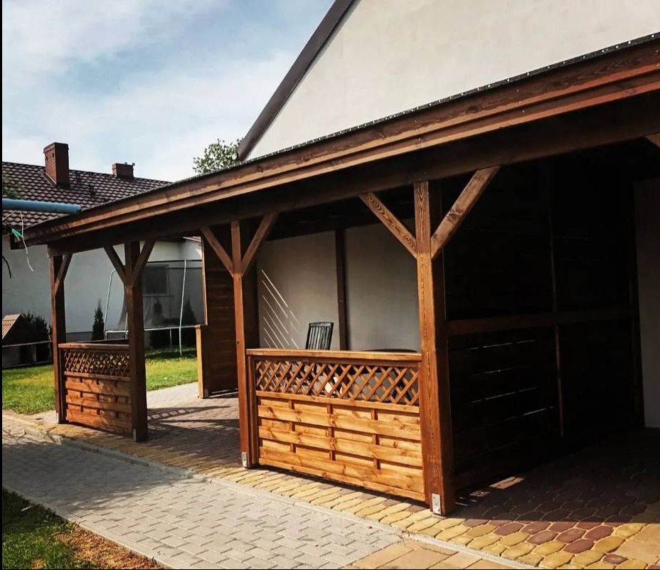 Zadaszona, drewniana konstrukcja z ażurowymi panelami, przylegająca do budynku. Widoczny fragment chodnika z kostki brukowej i trawnik. Umiarkowane oświetlenie.
