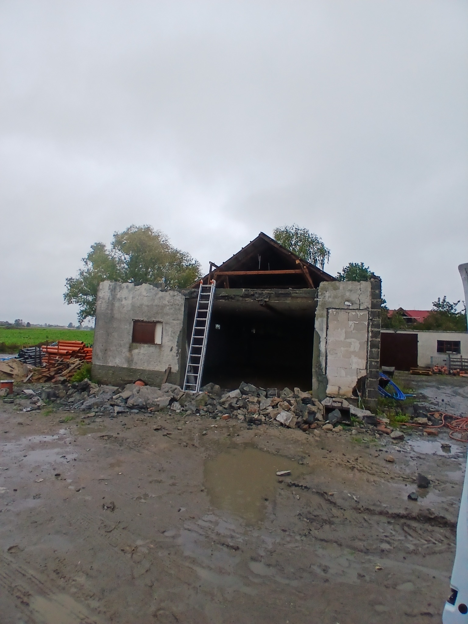 Zrujnowany budynek z drabiną opartą o ścianę, gruz na ziemi i widoczne elementy konstrukcyjne dachu. W tle zieleń i pochmurne niebo.