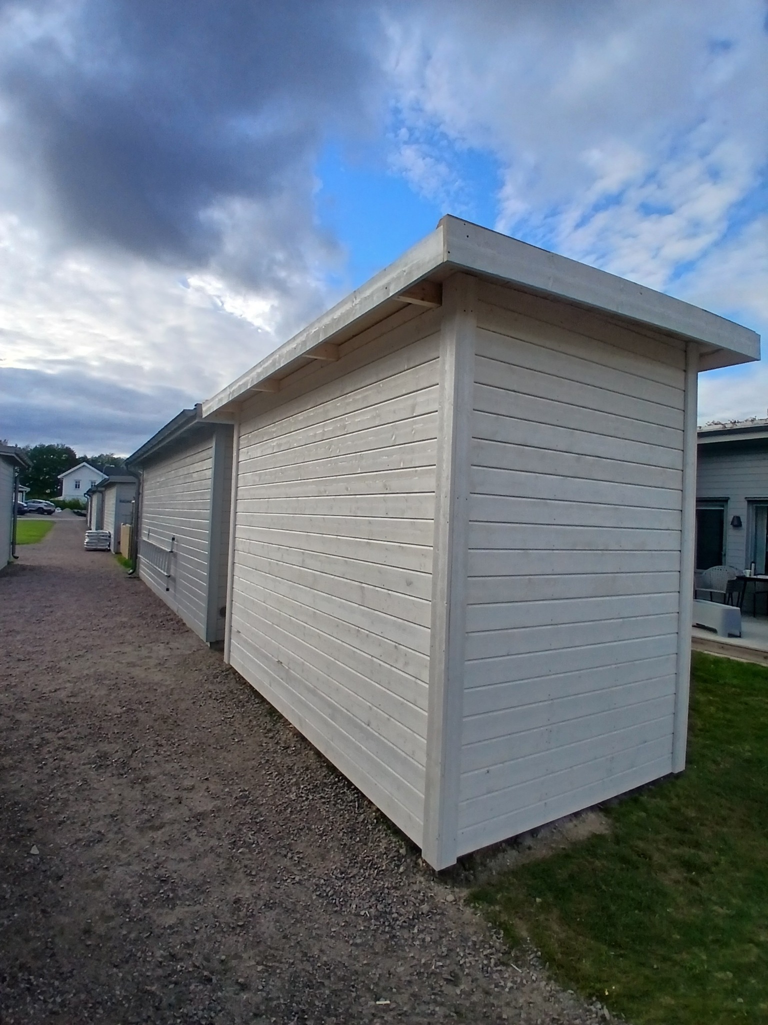 Biała, drewniana altana z płaskim dachem, widziana z boku na tle pochmurnego nieba i innych budynków. Widoczna faktura drewna i otoczenie.