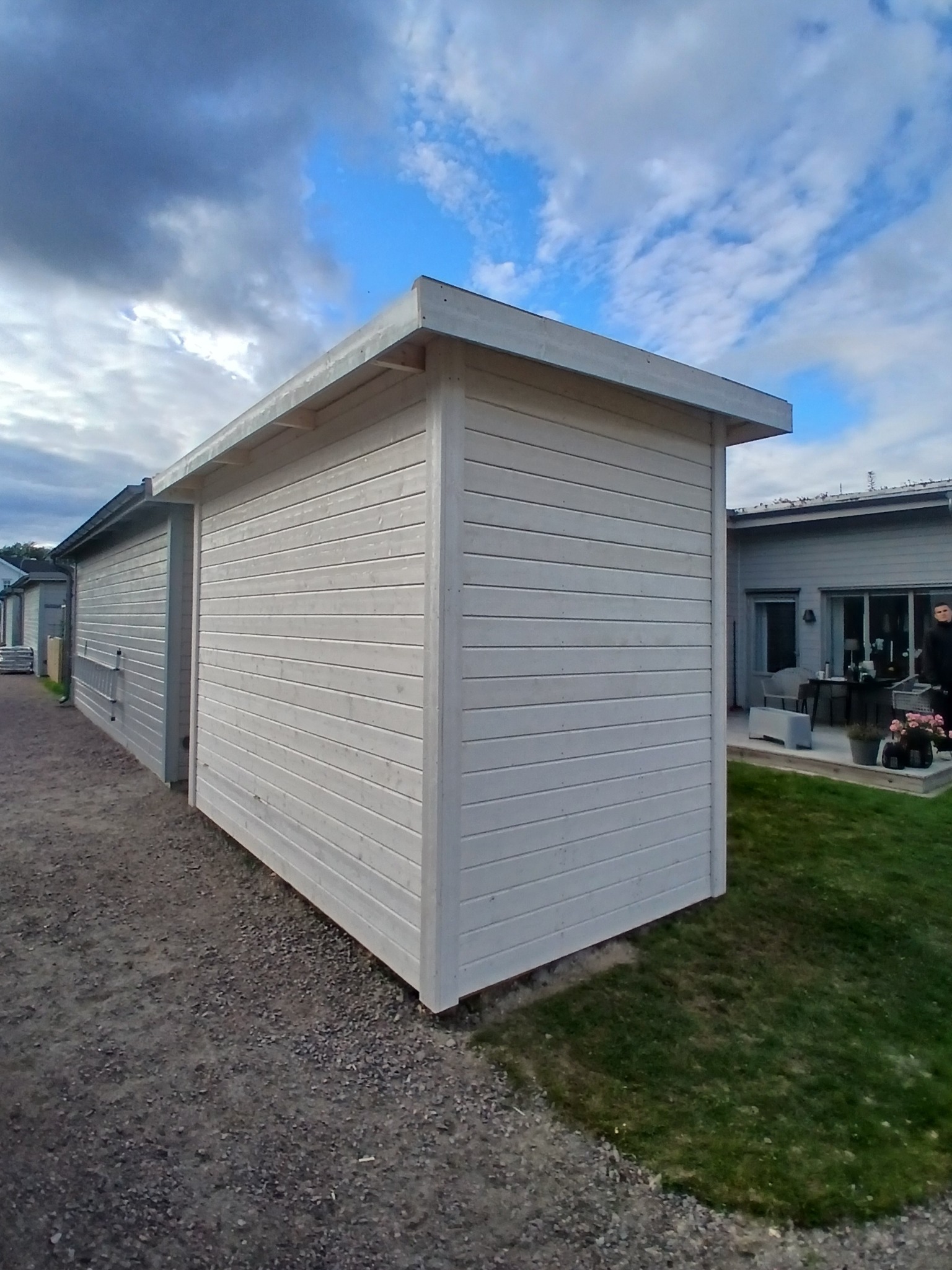 Biała, drewniana altana z płaskim dachem, widoczna z boku na tle pochmurnego nieba i zielonego trawnika. Minimalistyczny design, poziome deski.