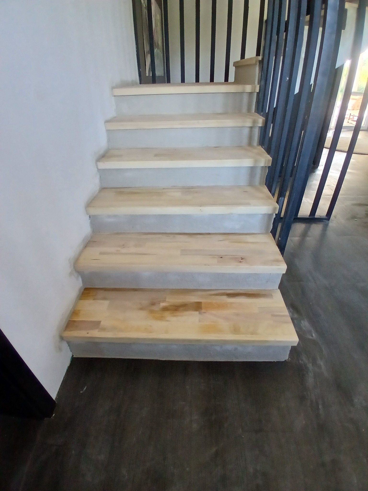 Jasne, drewniane schody z szarą, betonową podstawą i czarną, metalową balustradą w nowoczesnym wnętrzu. Widok z dołu, ukazujący detale wykończenia.