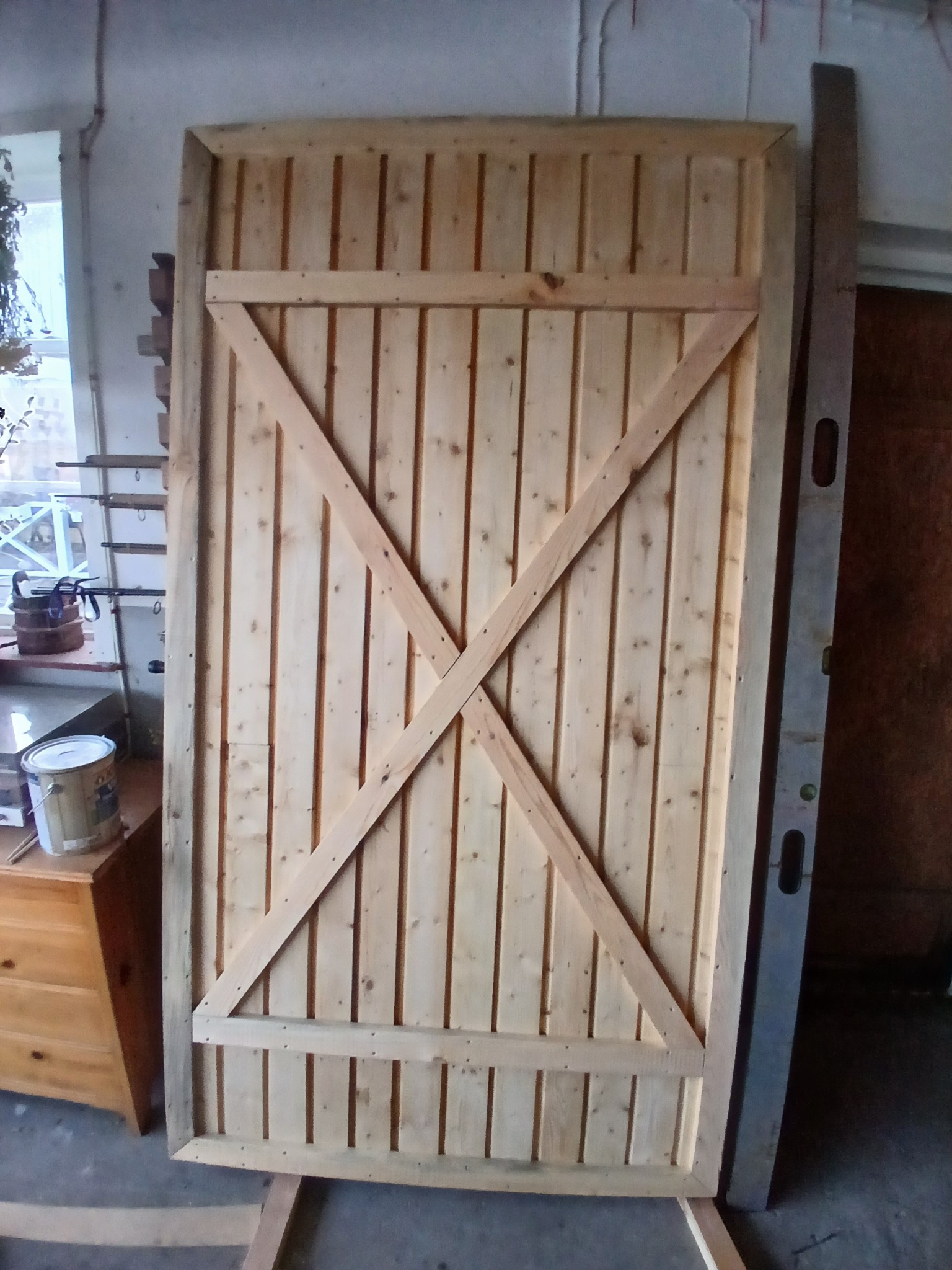 Surowe drewniane drzwi z ukośnymi wzmocnieniami w kształcie litery X, oparte o podłoże, obok poziomica i komoda z farbą, w tle okno i ściana.