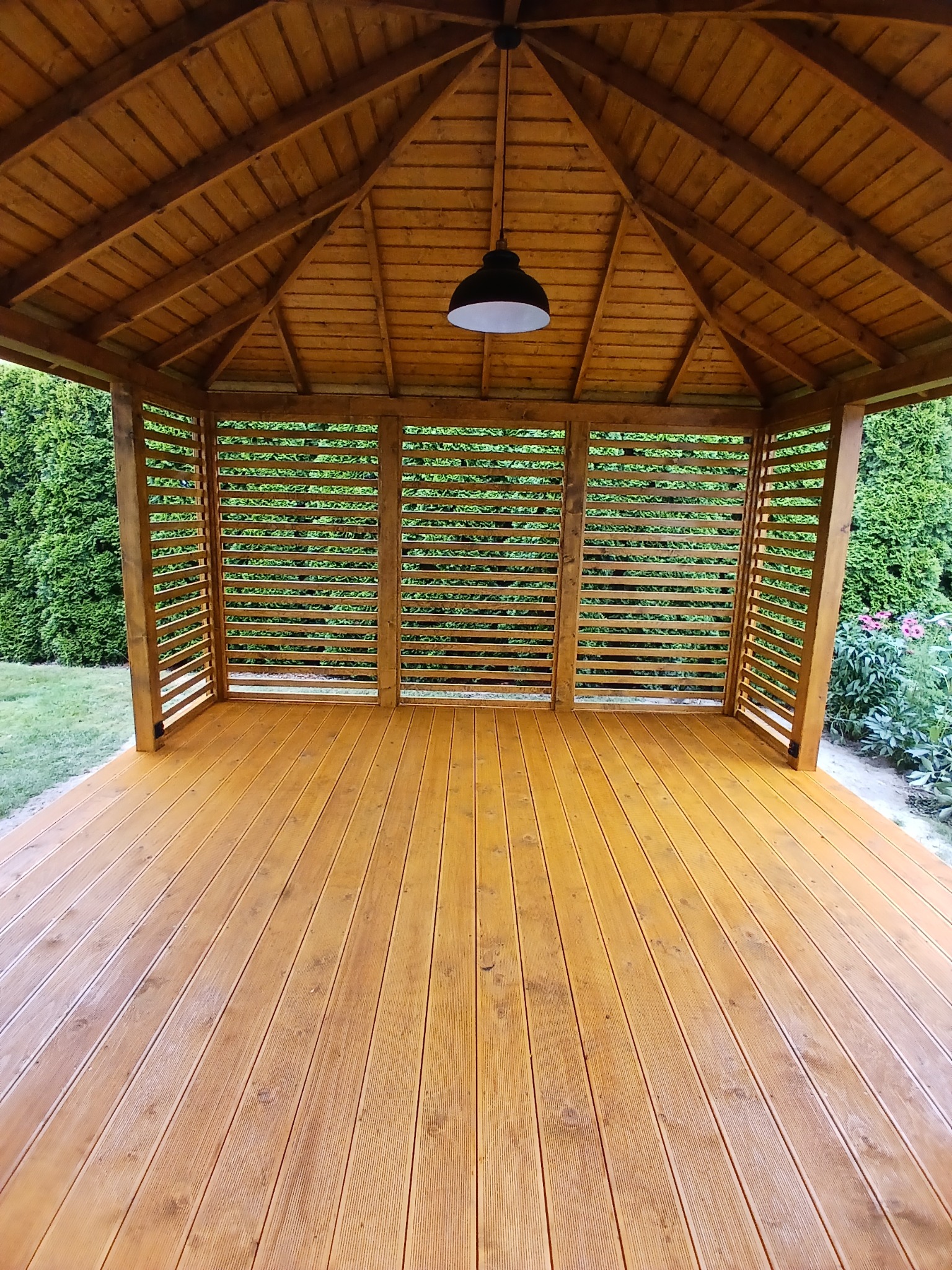 Drewniana altana z ażurowymi ścianami i podłogą z desek, oświetlona wiszącą lampą. Widok z wnętrza na zieleń ogrodu. Charakterystyczna konstrukcja dachu.