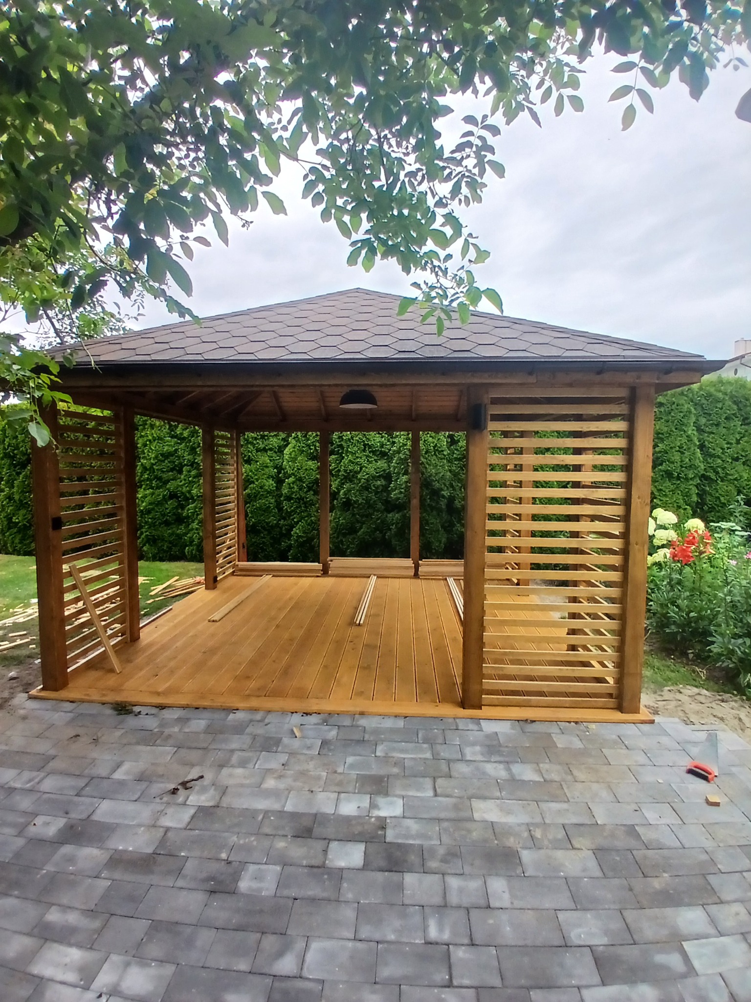 Drewniana altana z brązowym dachem na tle zieleni, częściowo wykończona, z widocznymi elementami konstrukcyjnymi i narzędziami na kostce brukowej.